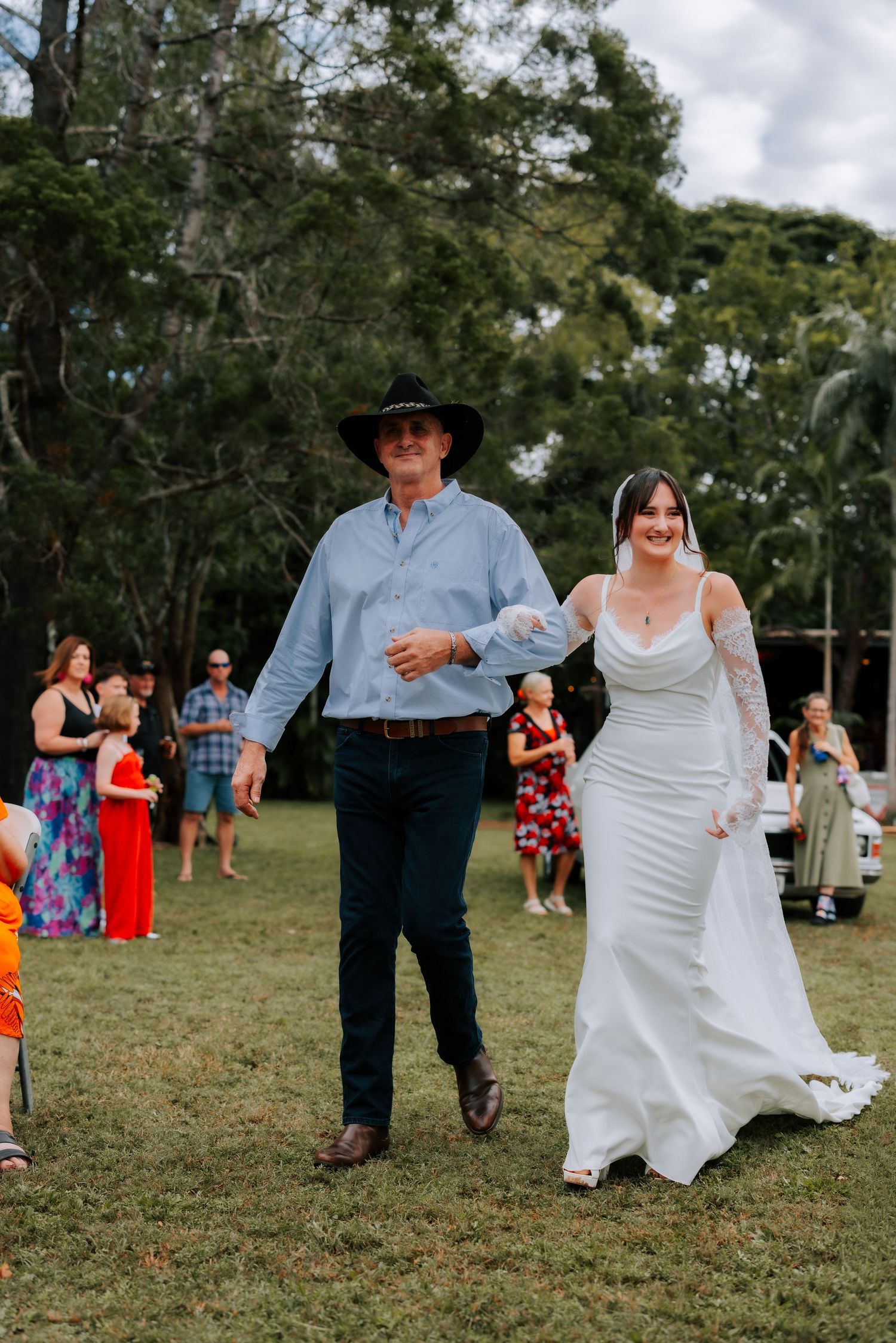 Bride in white lace gown walks with older man in cowboy hat down outdoor aisle.