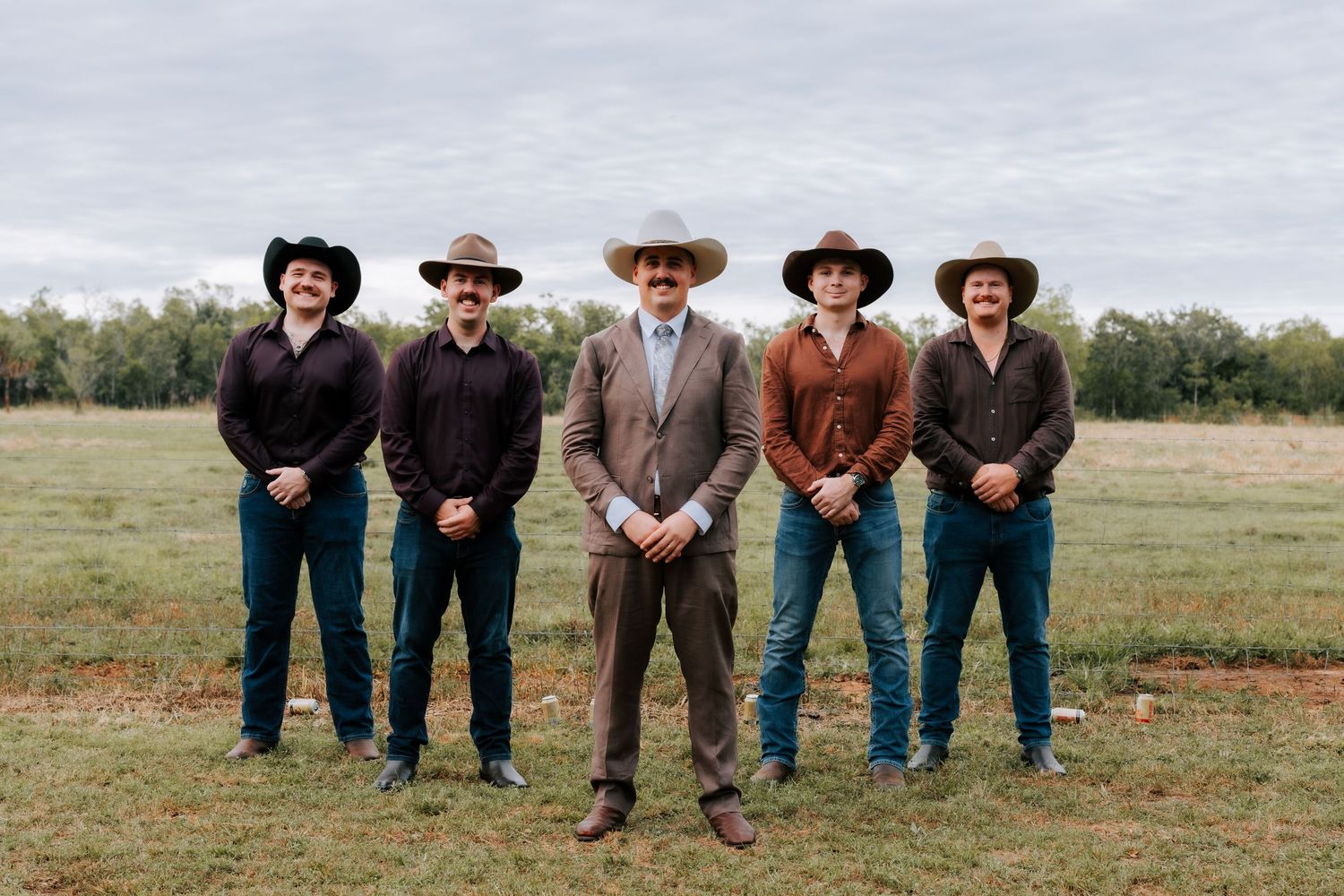 Five groomsmen wearing cowboy hats and brown suits posing in a green field.
