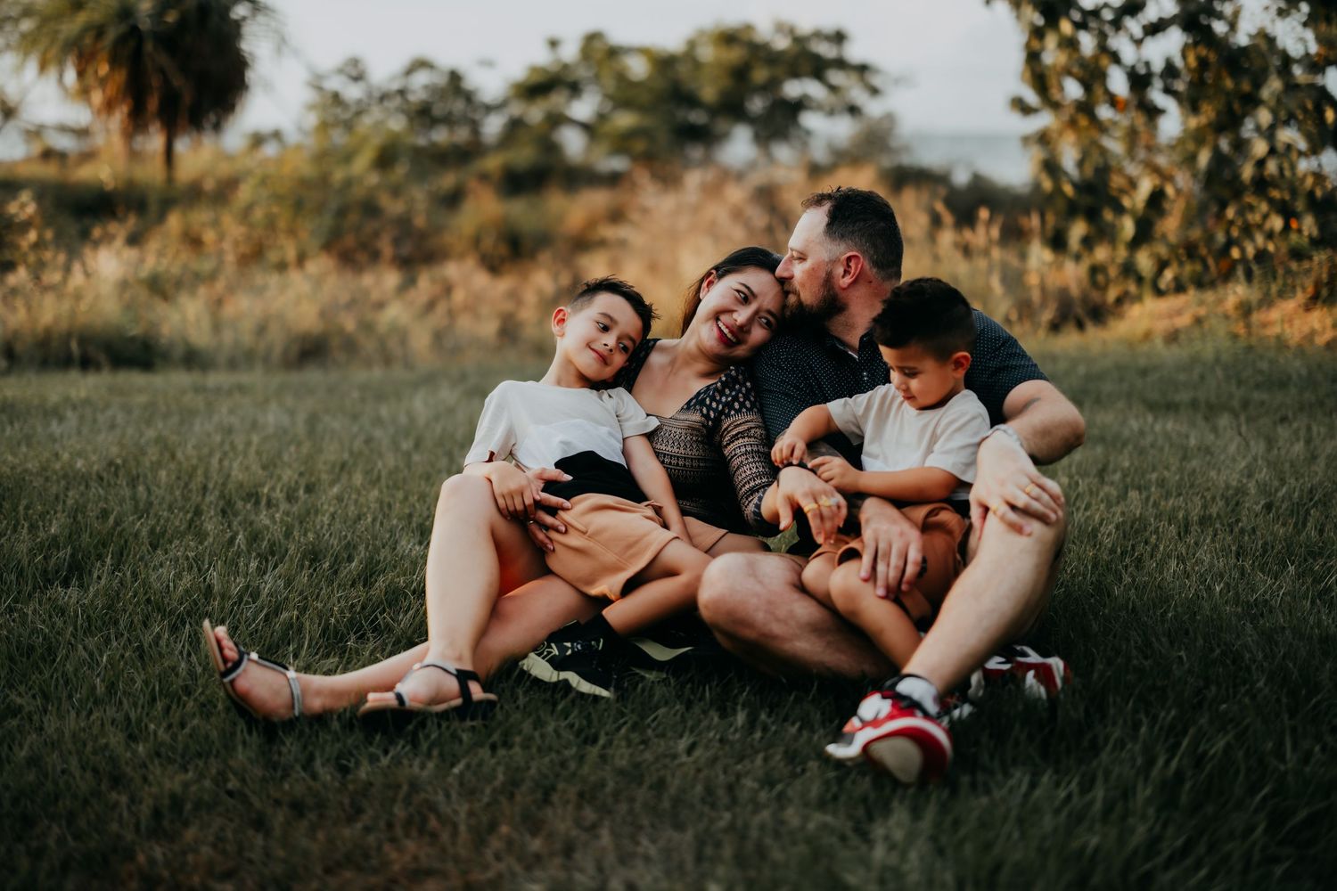 A loving family of four sits closely together on grass, sharing hugs and smiles during an outdoor photo session.