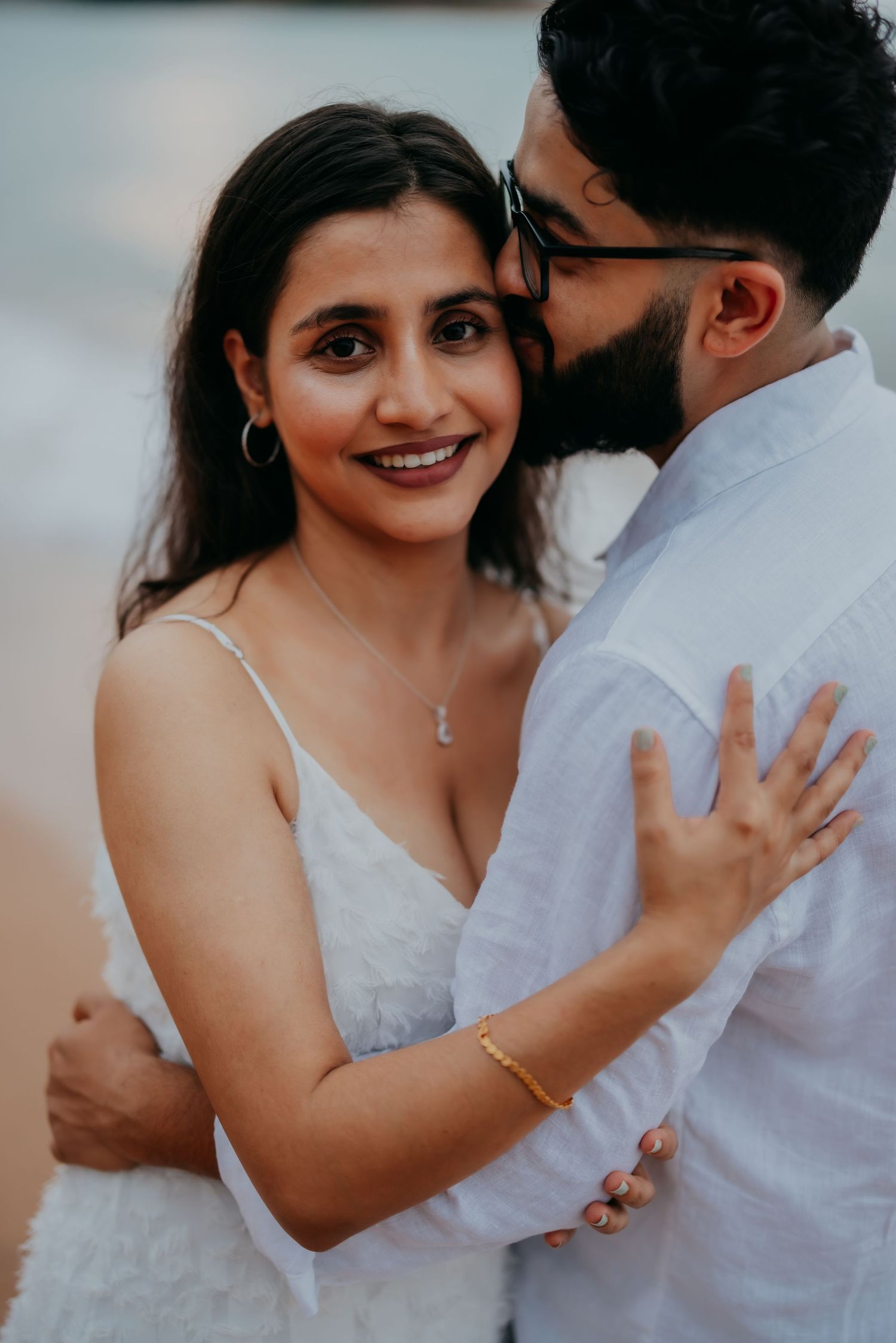 Bride in white dress smiling as groom kisses her cheek during romantic beach engagement photo session.
