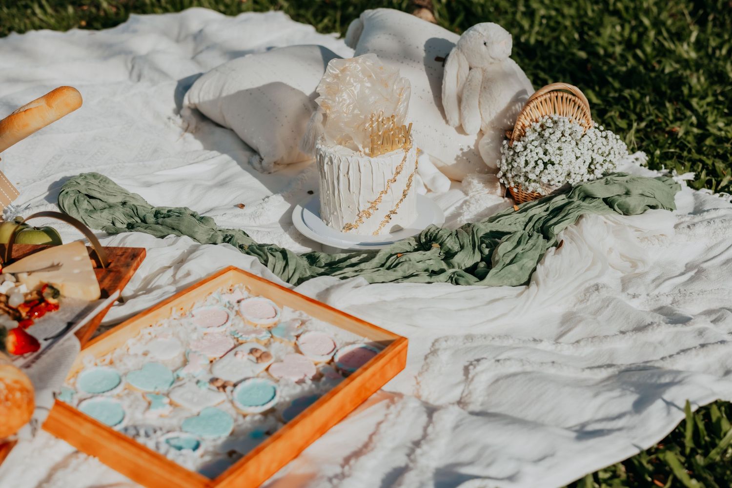 Elegant outdoor picnic setup with white cake, eucalyptus garland, cheese board, and decorative tray on white blanket.