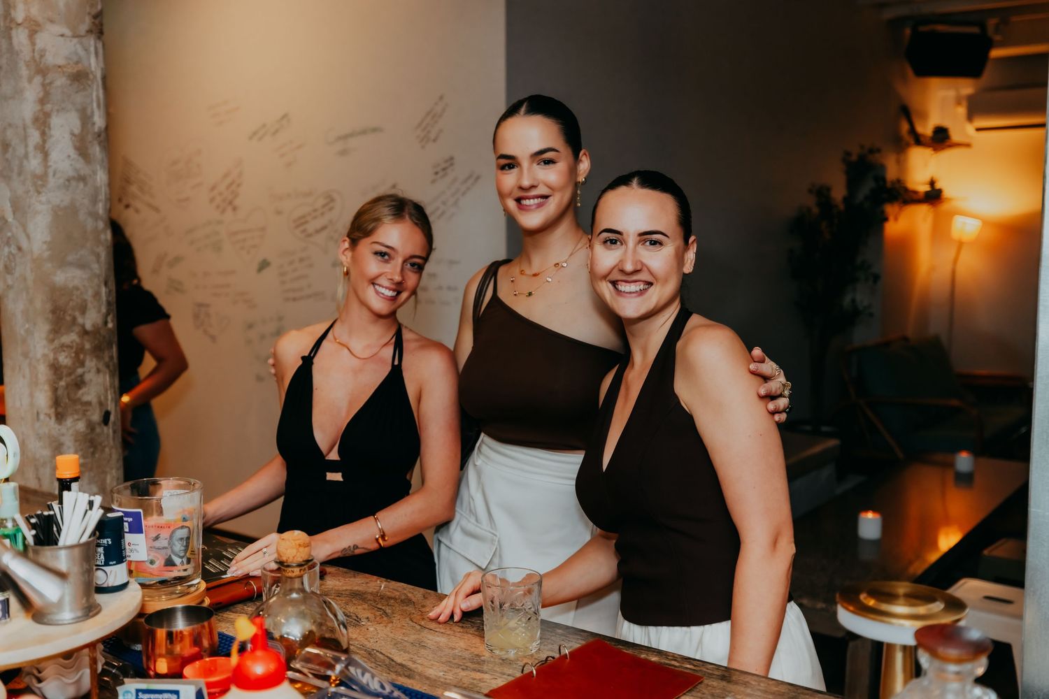 Three smiling women in black outfits posing together at a bar counter in a stylish indoor venue.