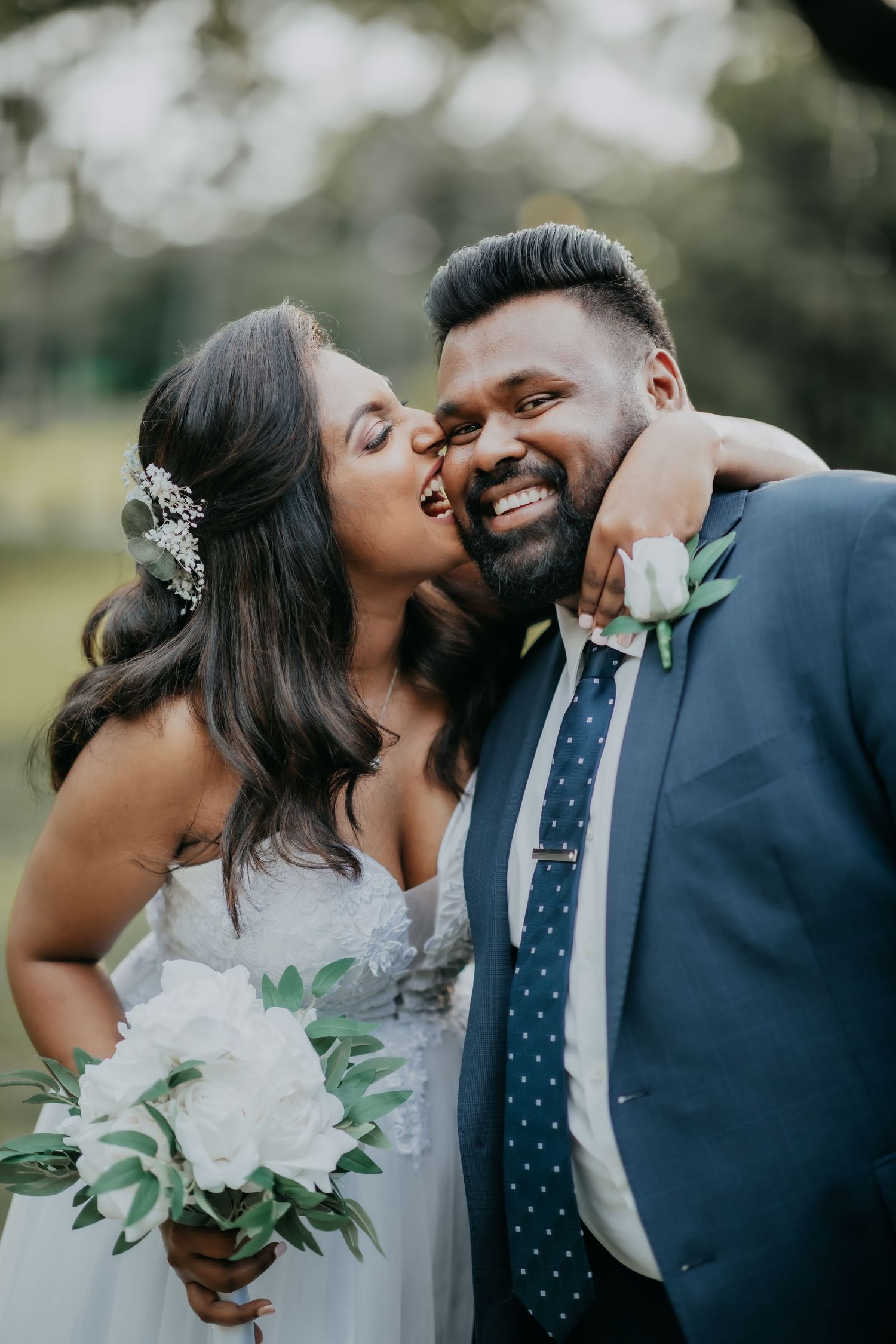 Joyful bride kissing groom on cheek outdoors, groom wearing navy suit with white boutonniere.