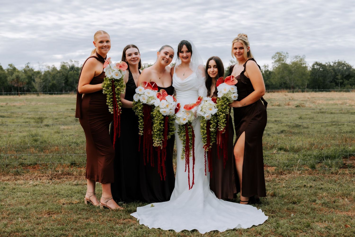 Bride in white gown with four bridesmaids in dark dresses holding cascading bouquets outdoors.