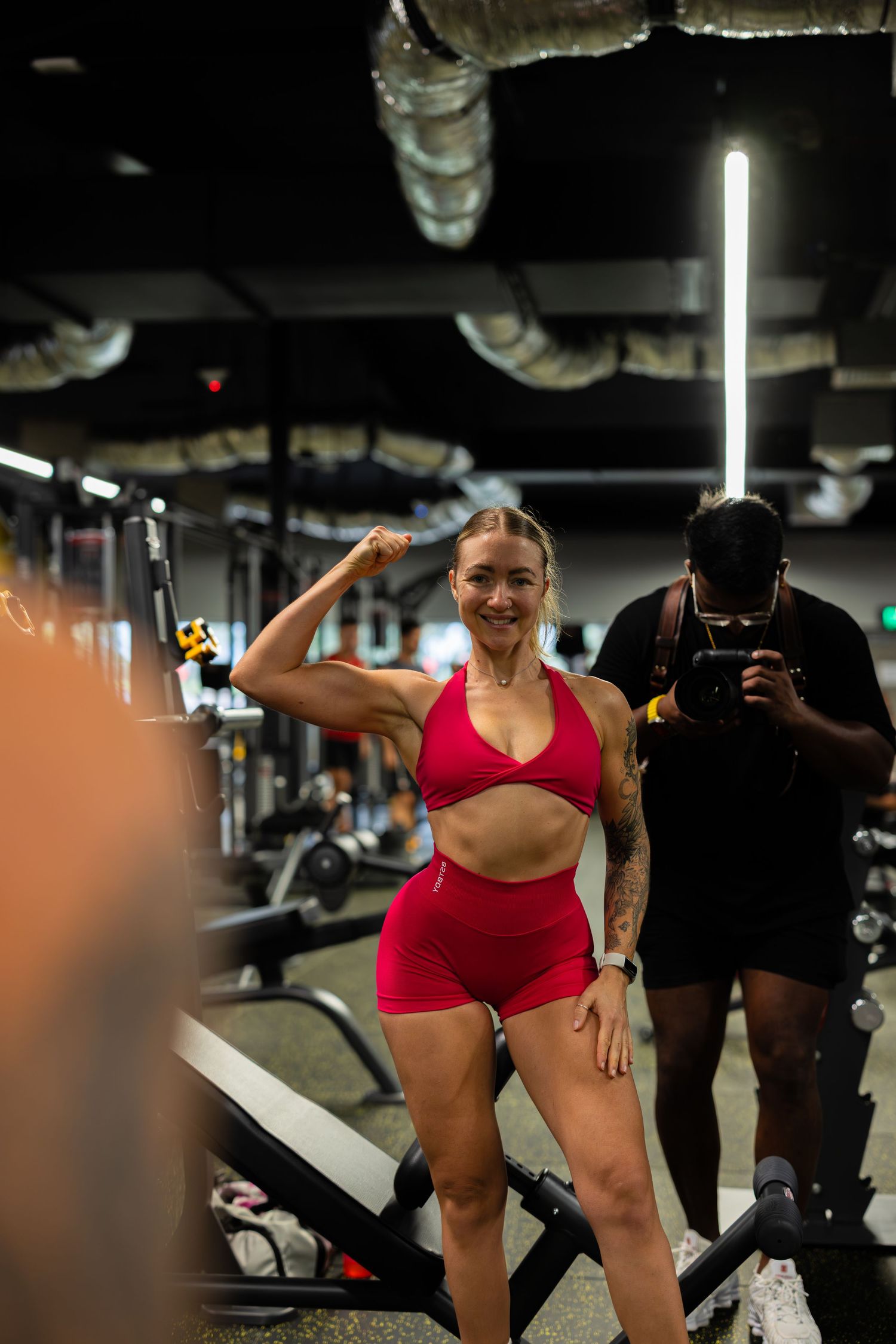 Fit blonde woman in red gym outfit flexing her bicep muscle while smiling in a gym.