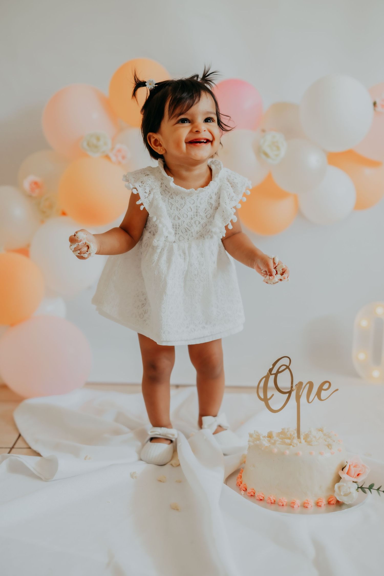 Happy one-year-old girl in white dress laughing during cake smash with peach balloon garland and One cake topper.