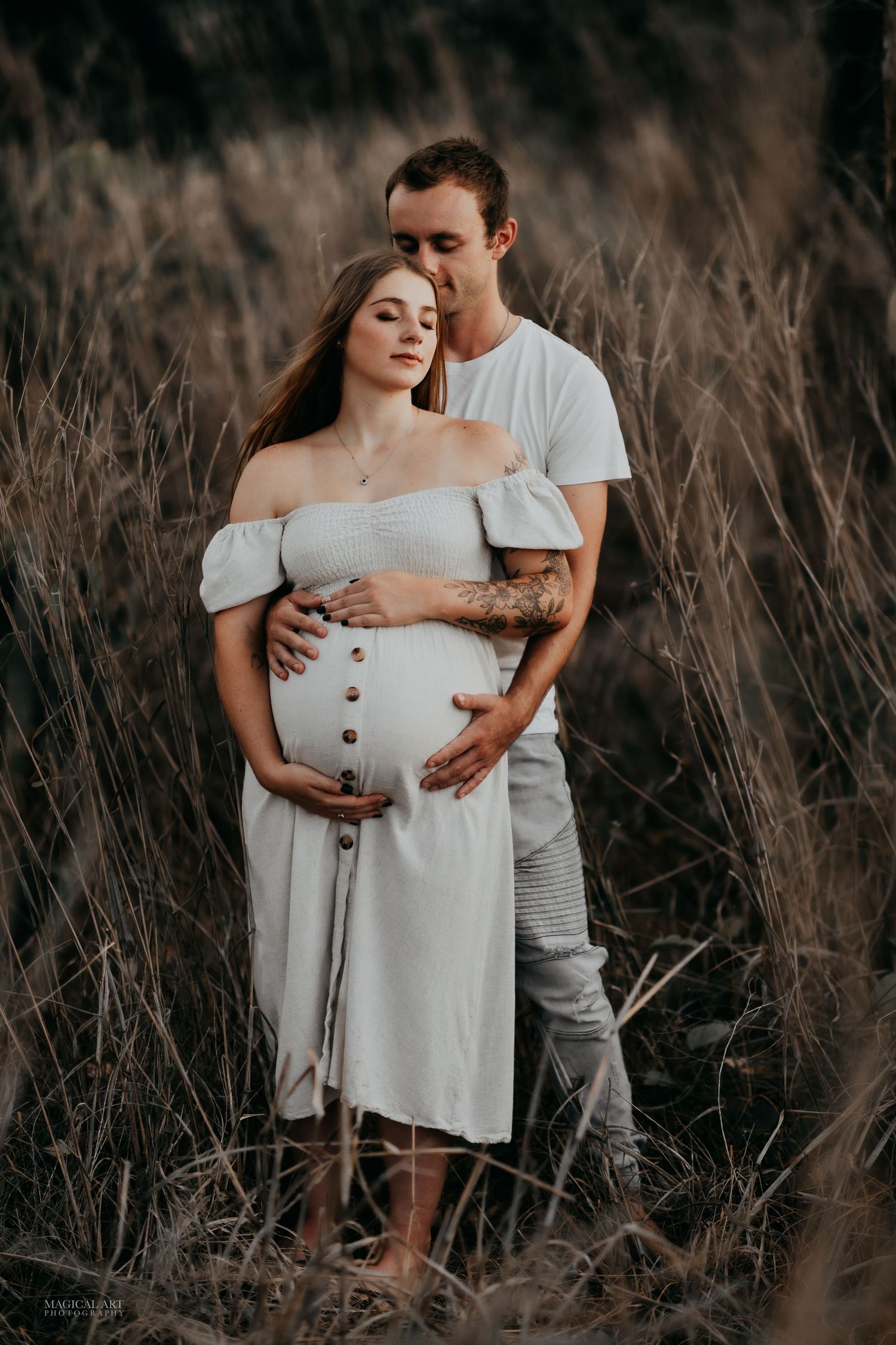 Couple embracing in tall grass at sunset, pregnant woman in white dress.