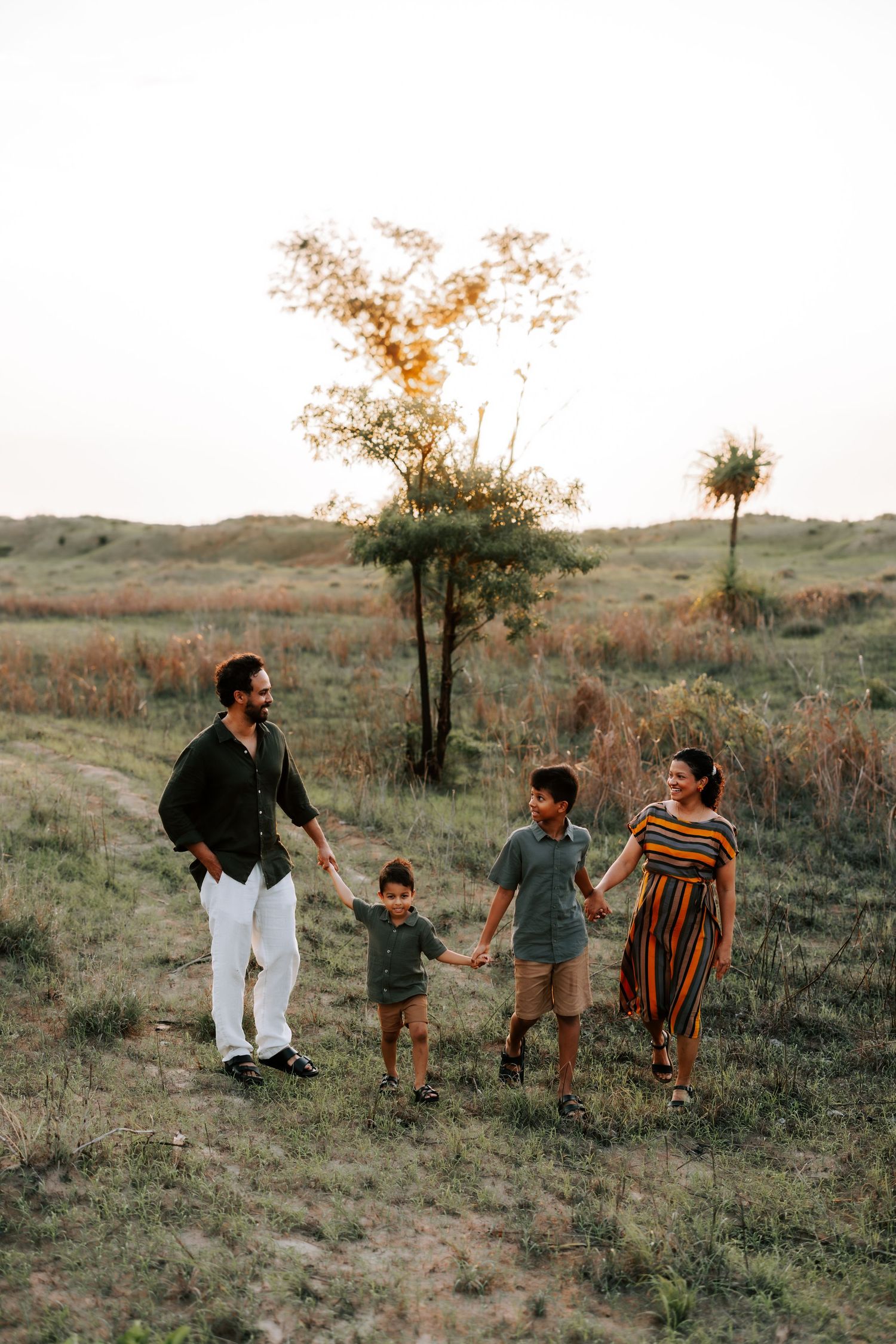 Happy family of four walking together through an open grassy field at sunset, holding hands and smiling.