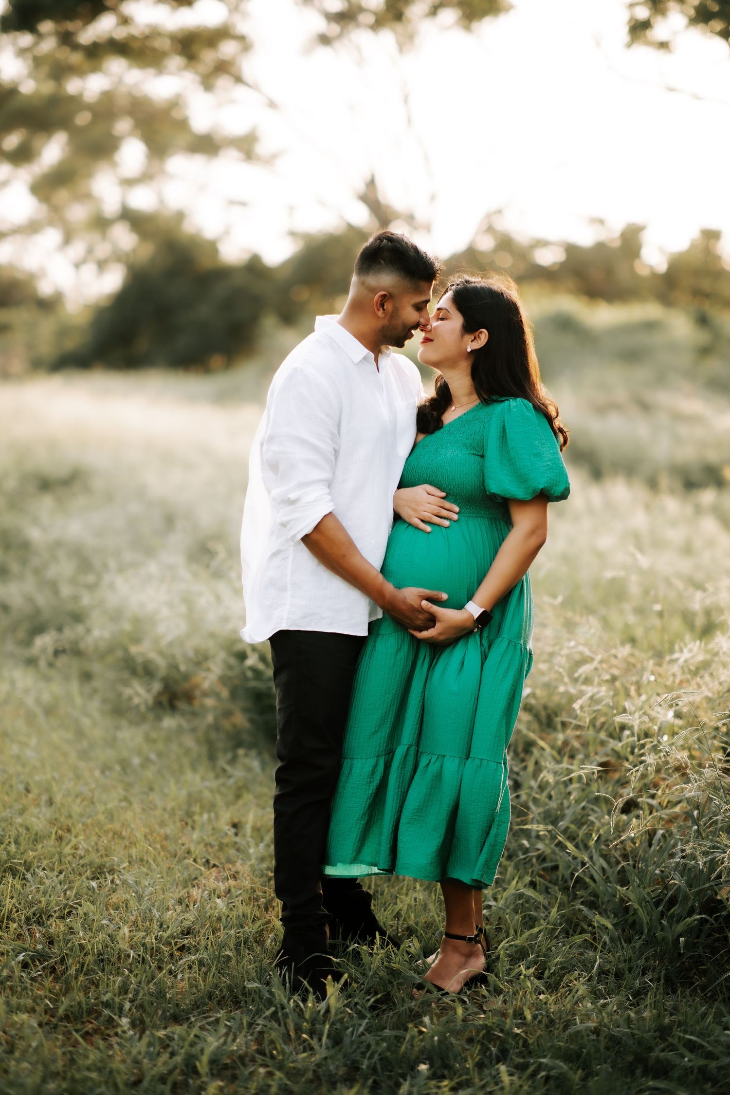 Couple sharing a kiss in a sunlit field, woman in green maternity dress.