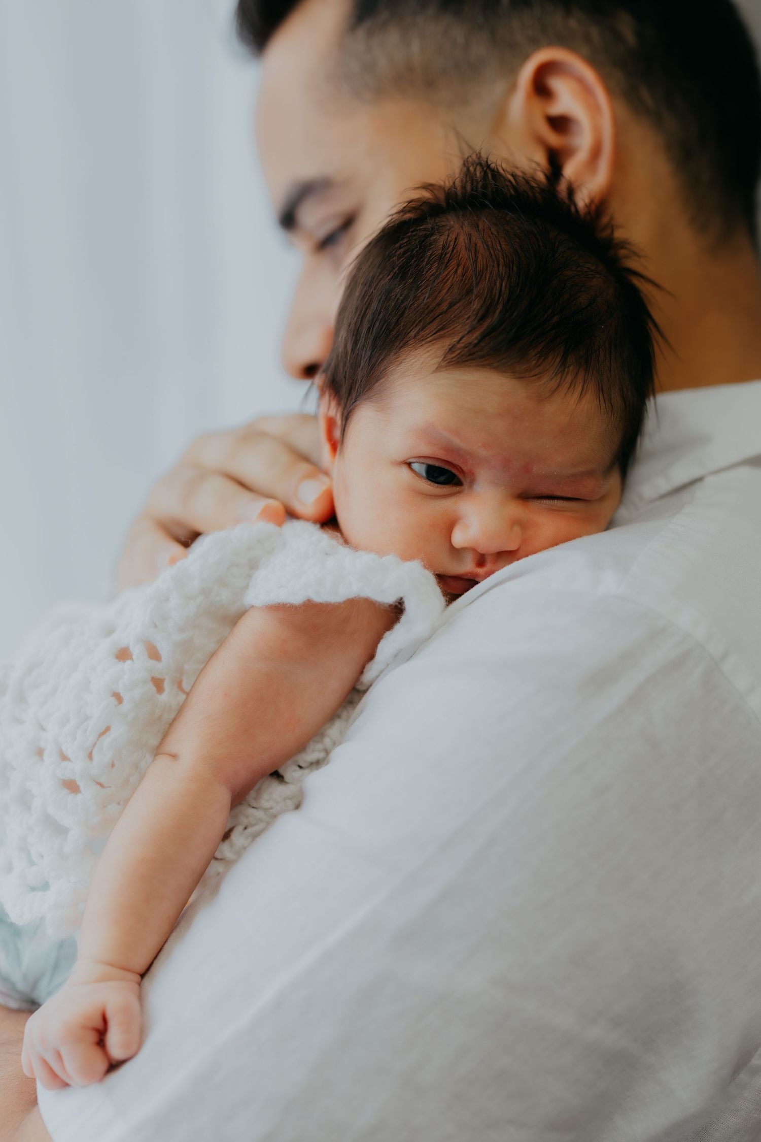 Father in white shirt gently cradling alert newborn baby against his chest.