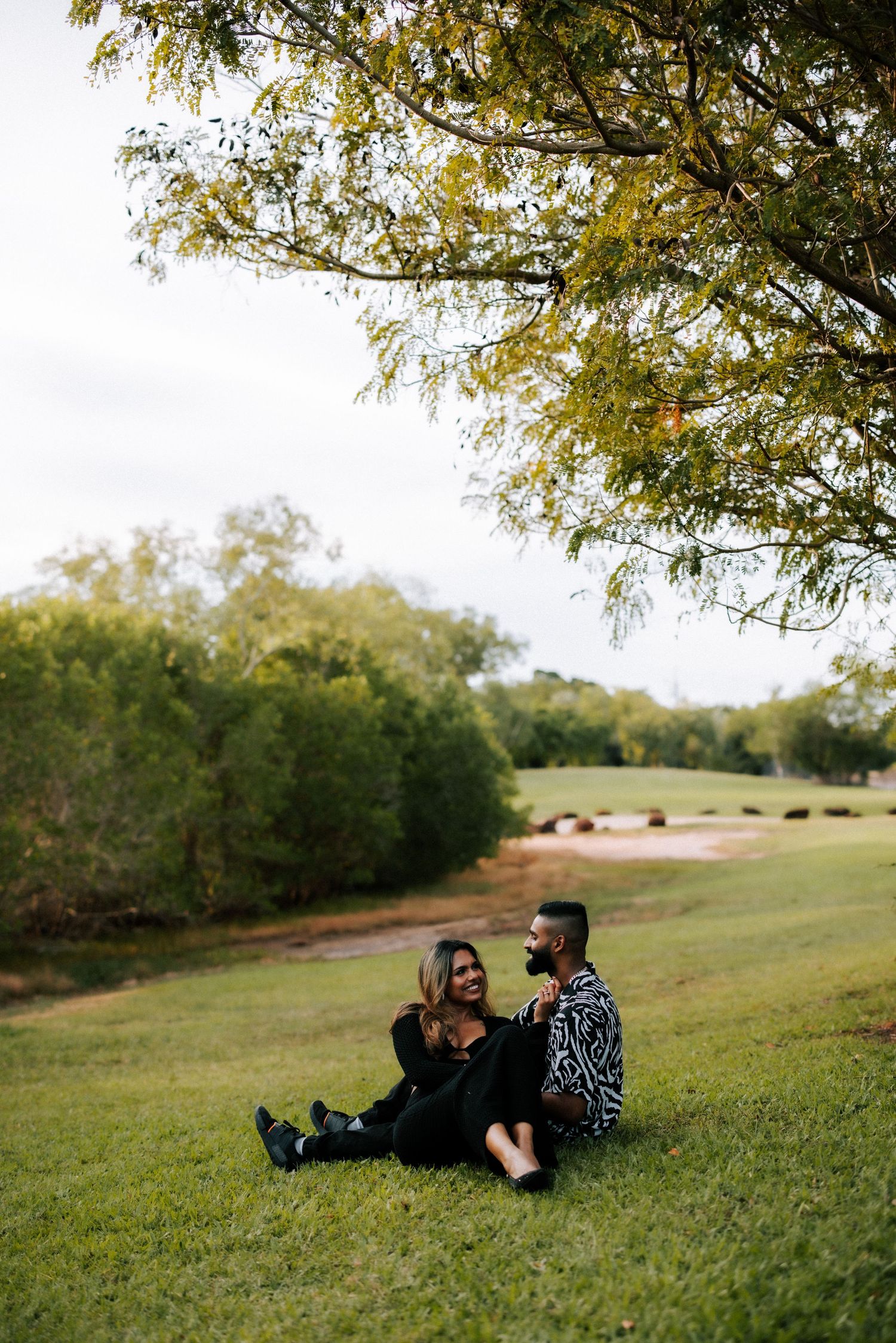 Couple sitting together on green grass under a large tree in a scenic outdoor park setting.