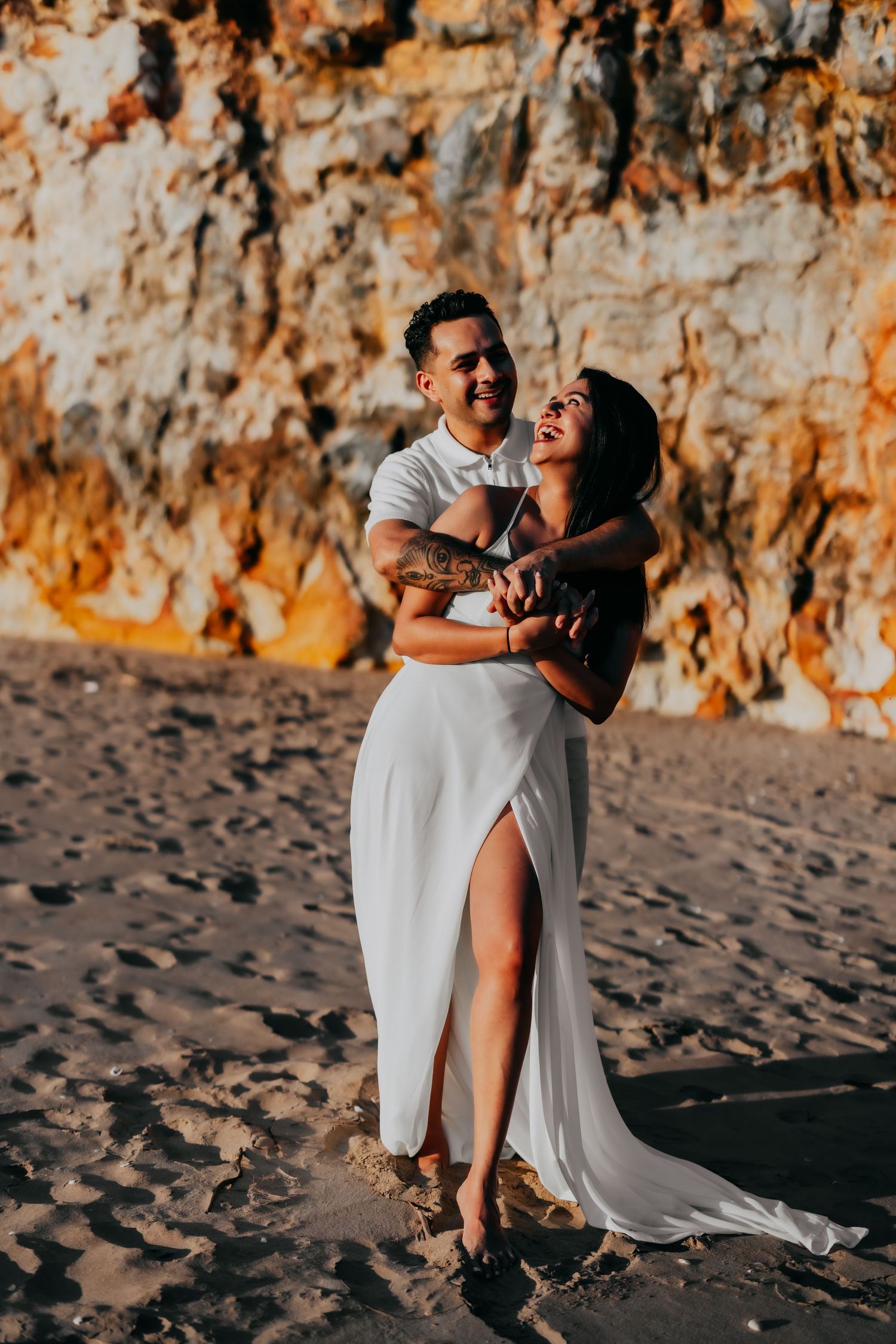 Joyful couple embracing and laughing on sandy beach with dramatic golden rocky cliffs in the background.