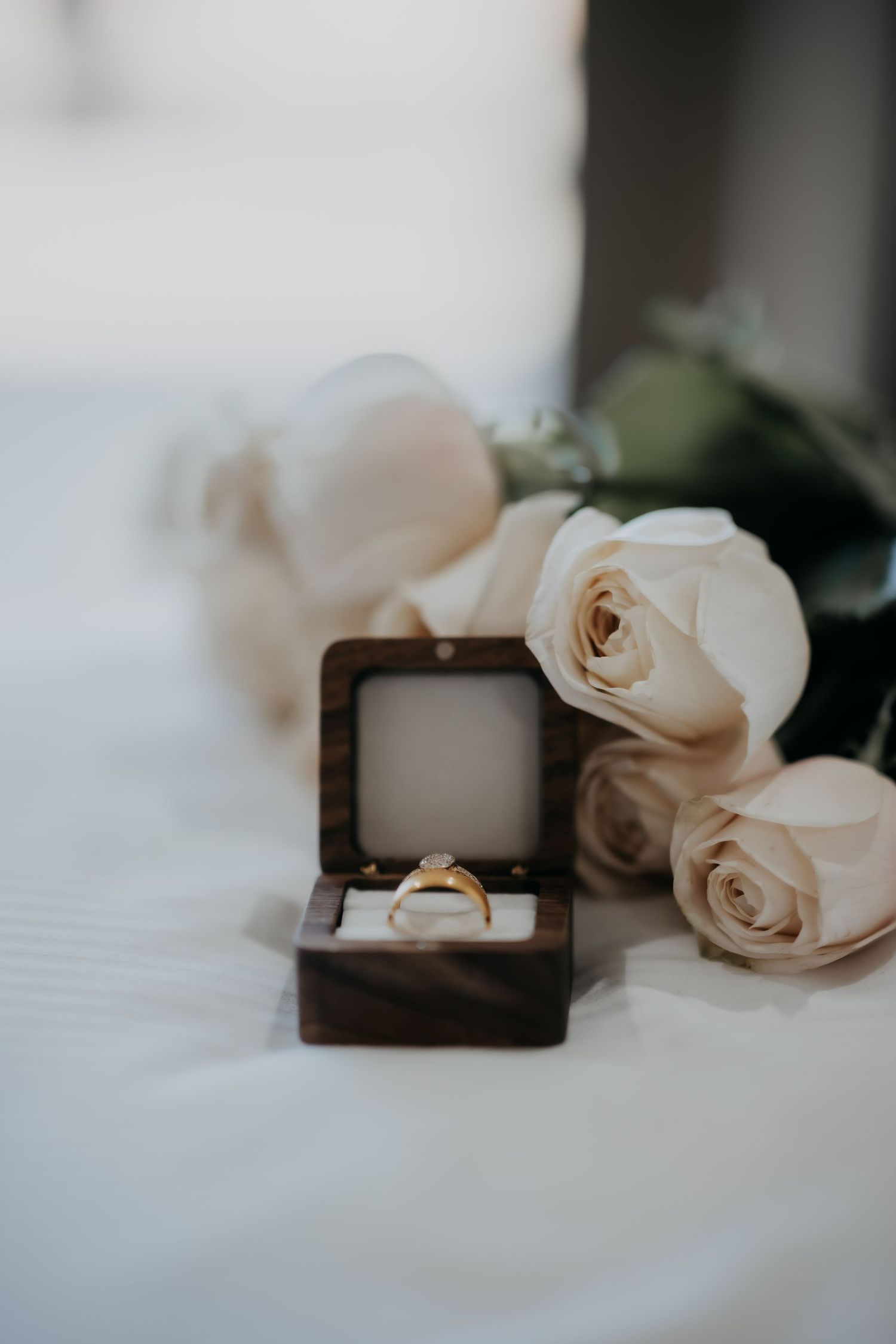 Gold diamond engagement ring in wooden box surrounded by white roses on white surface.