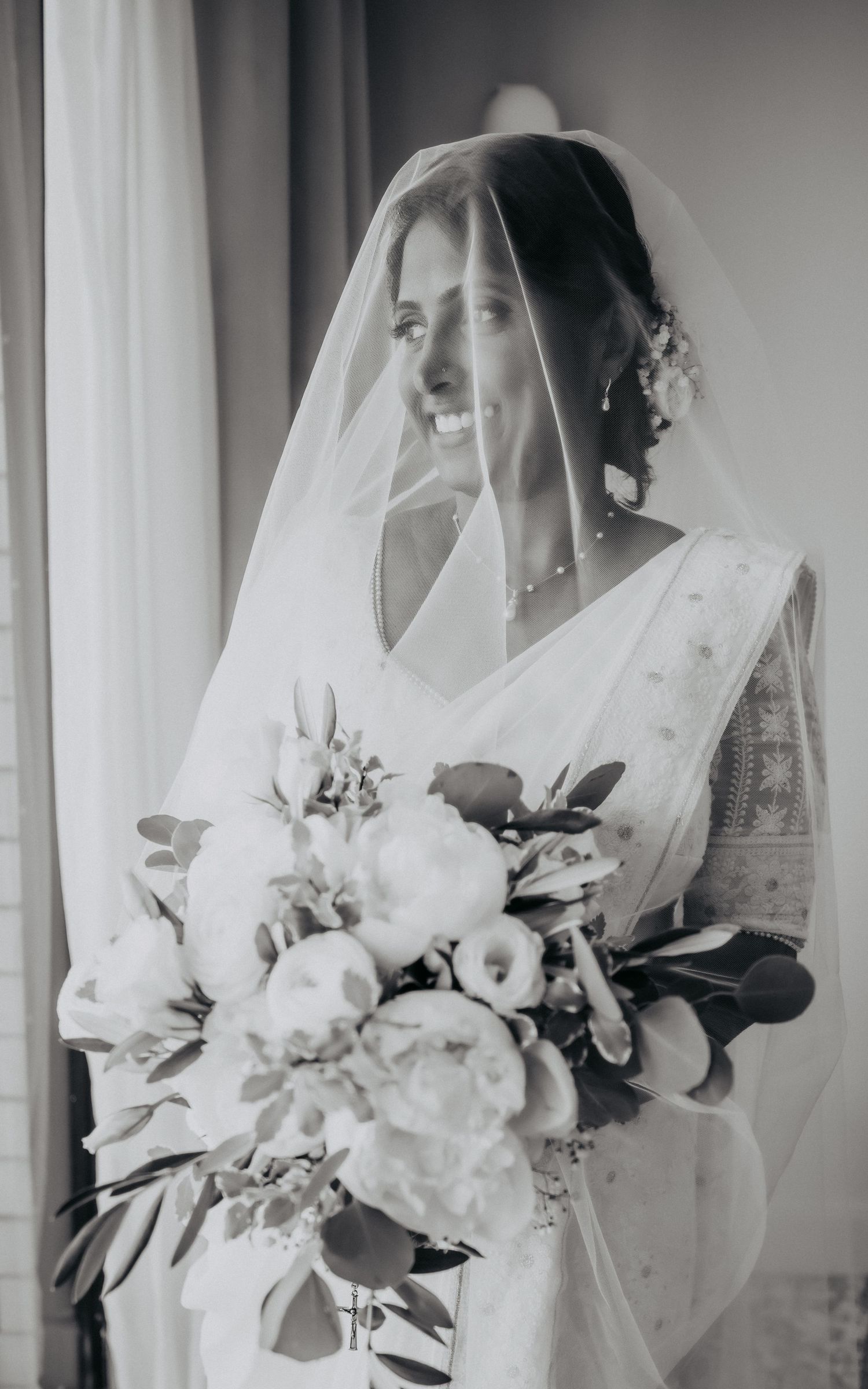 Black and white portrait of bride in white saree holding large floral bouquet with veil draped over face.