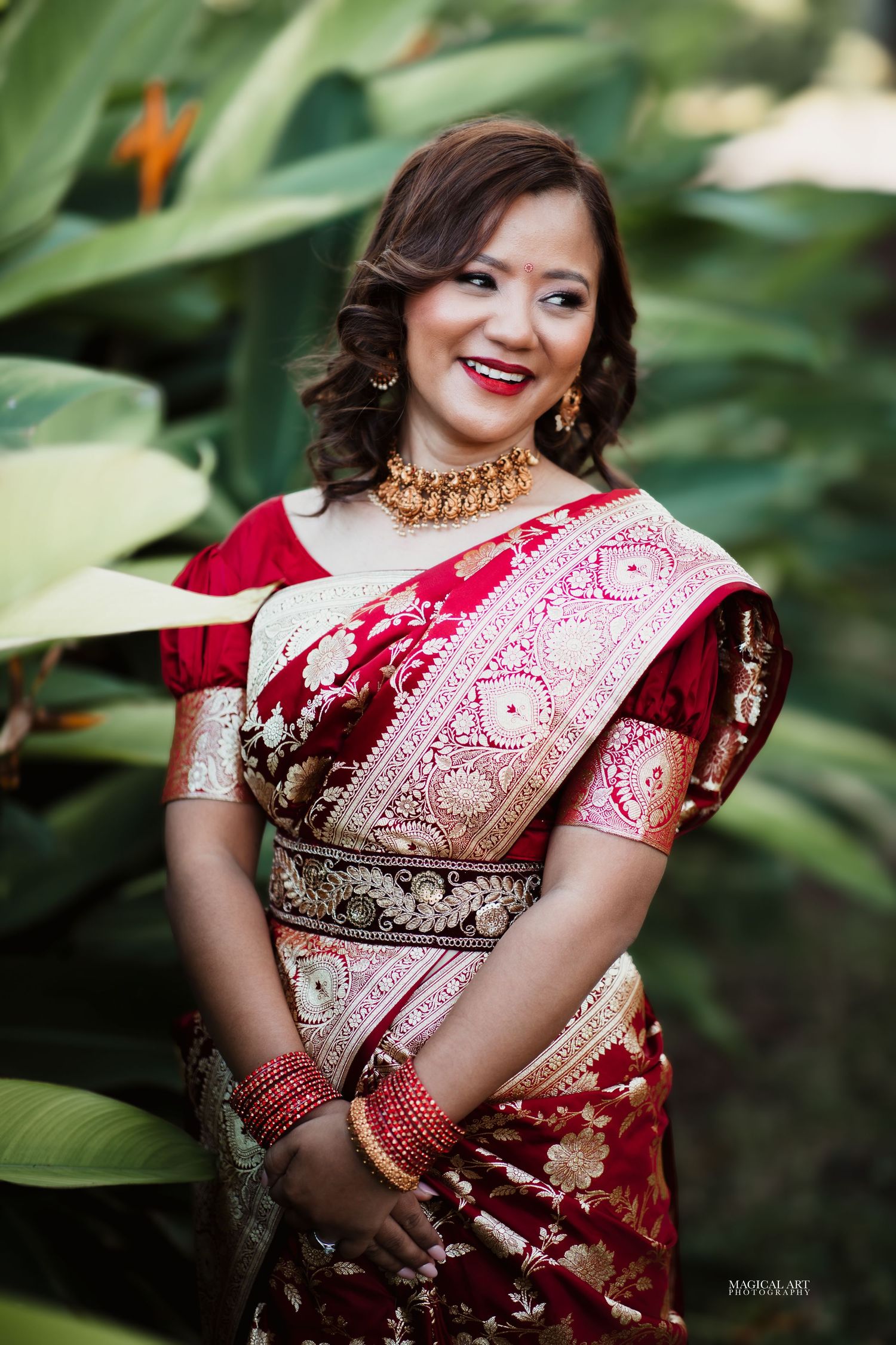 Smiling bride in red and gold Banarasi saree with gold choker necklace and bangles posing outdoors.
