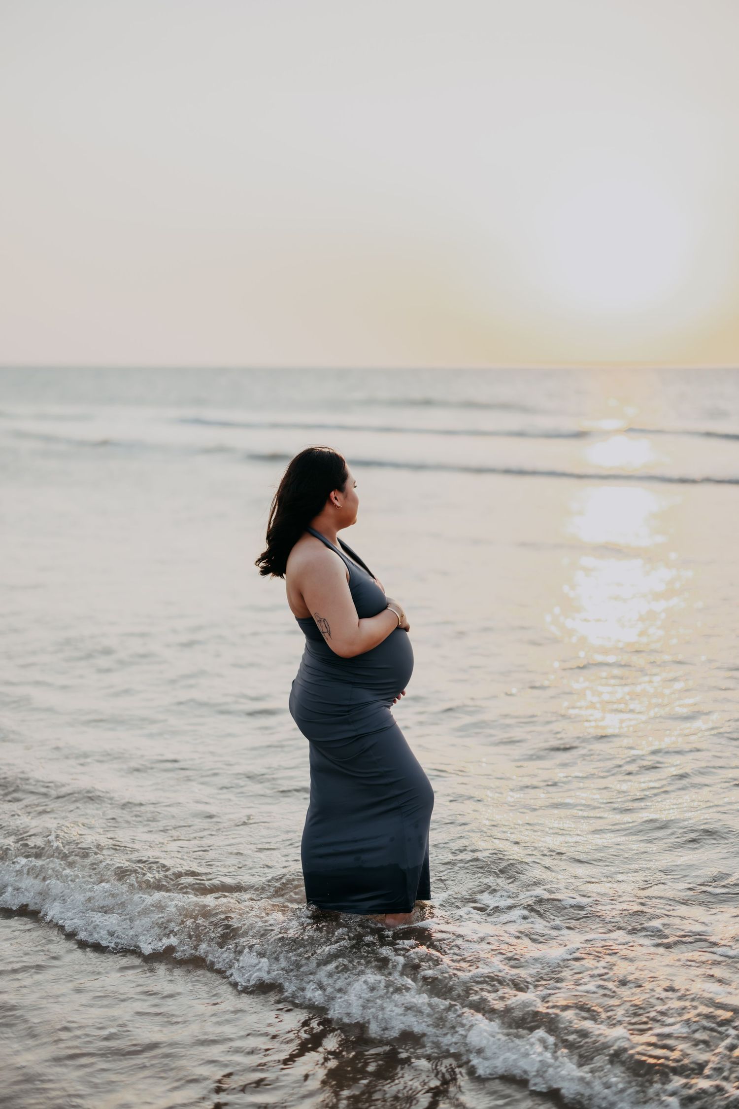Pregnant woman in grey dress standing in ocean waves at sunset holding her belly.