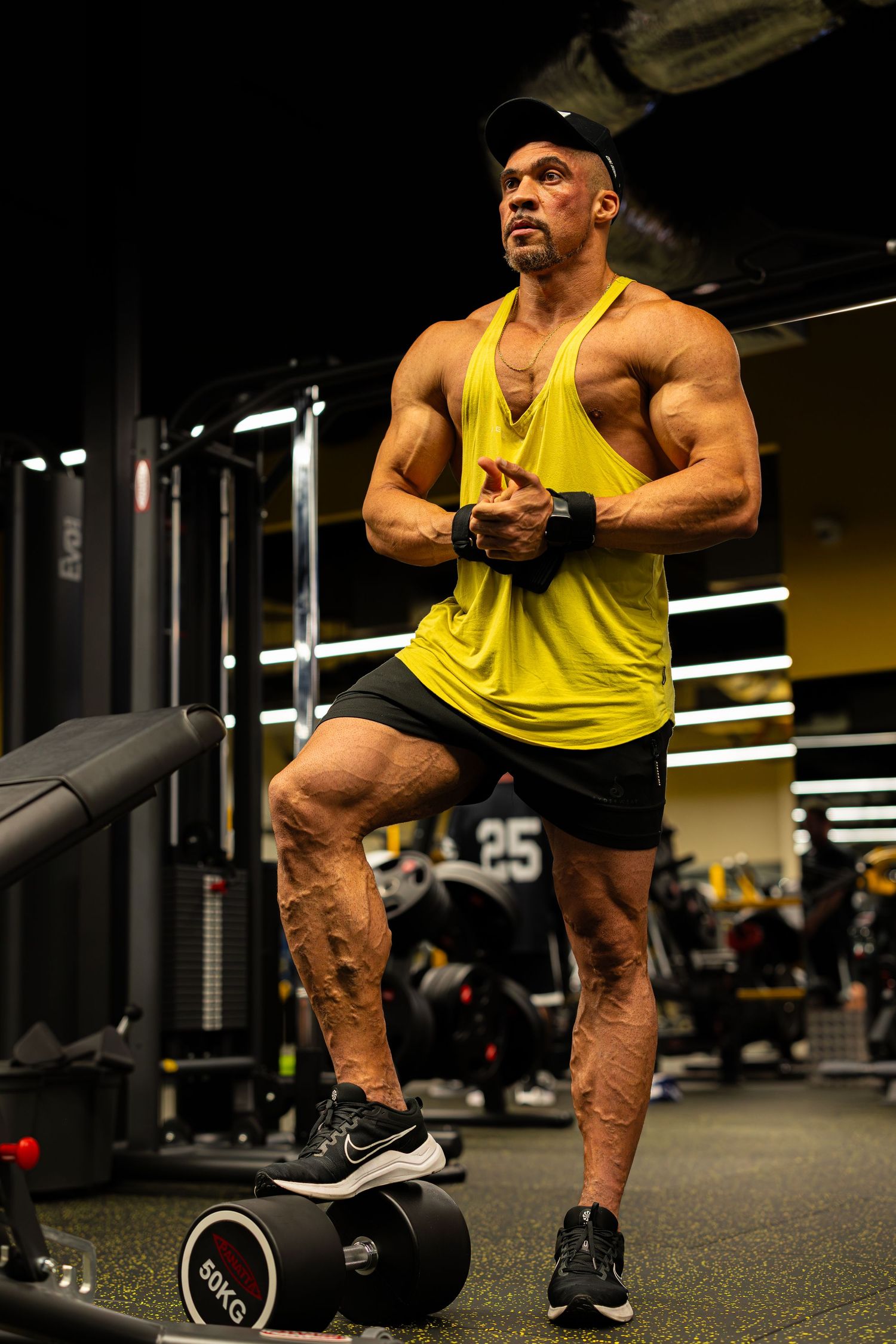 Muscular bodybuilder in yellow tank top and black shorts posing next to 80kg dumbbells in gym.