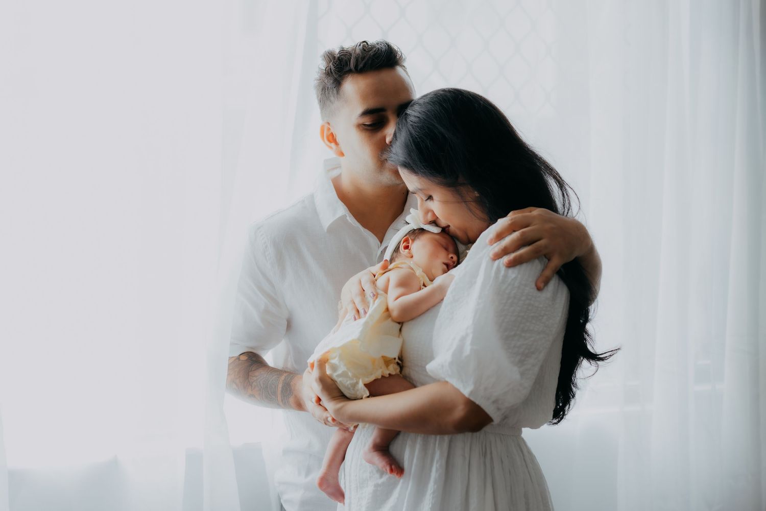 Parents in white outfits tenderly holding and kissing their newborn baby by a window.
