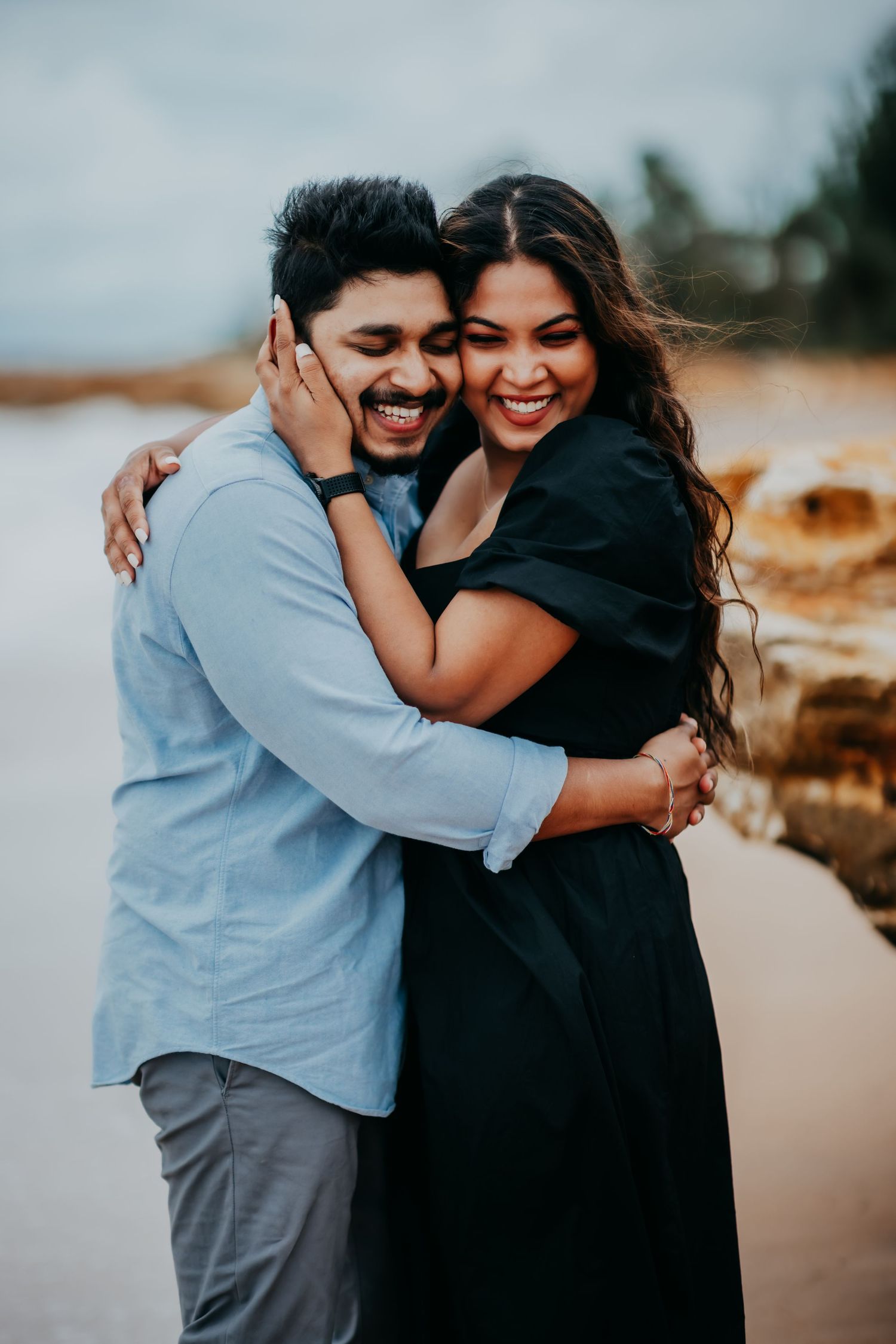 Laughing couple embracing on a rocky beach, woman in black dress hugging man in light blue shirt.