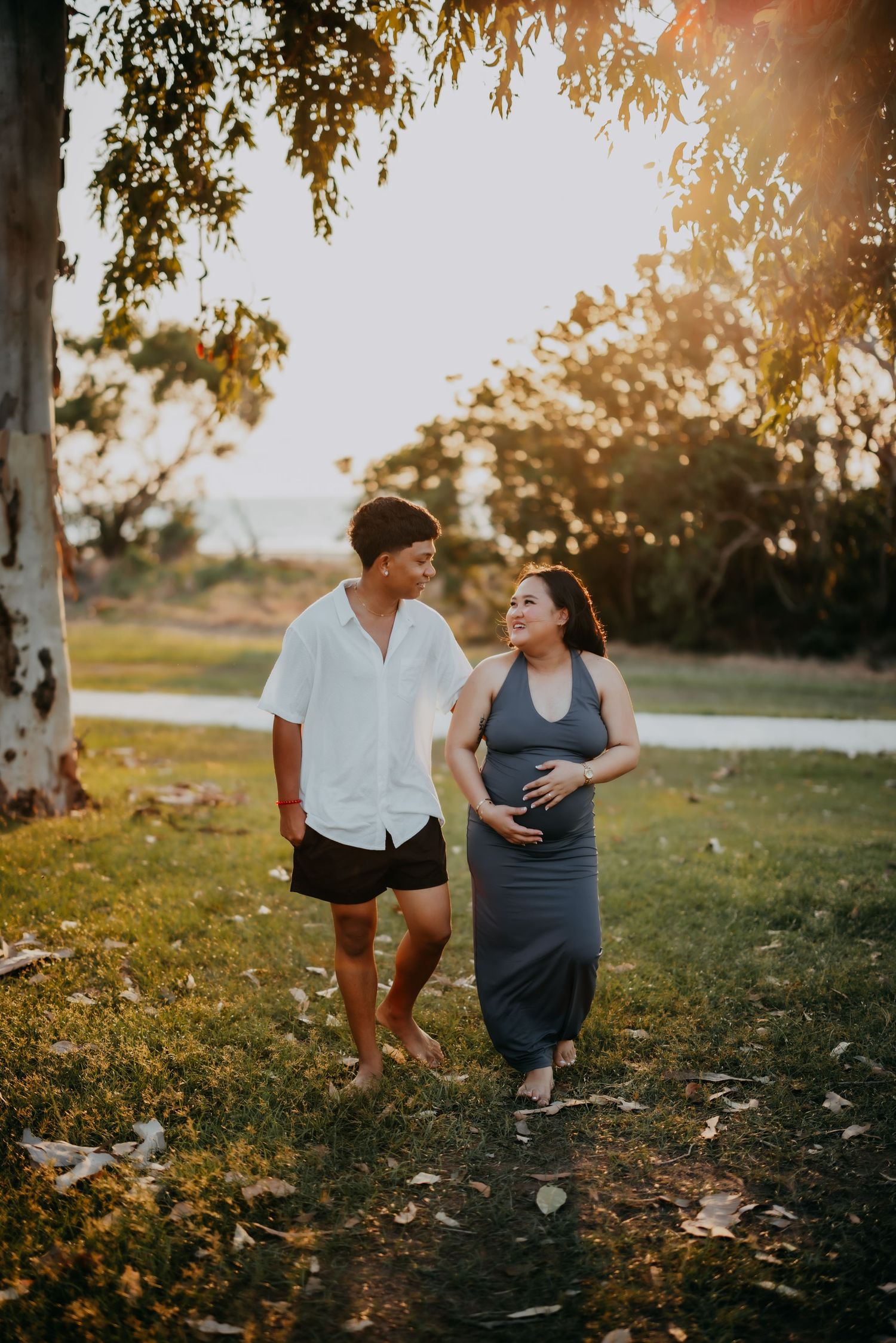 Couple walking together at golden hour, pregnant woman in grey dress holding her belly.