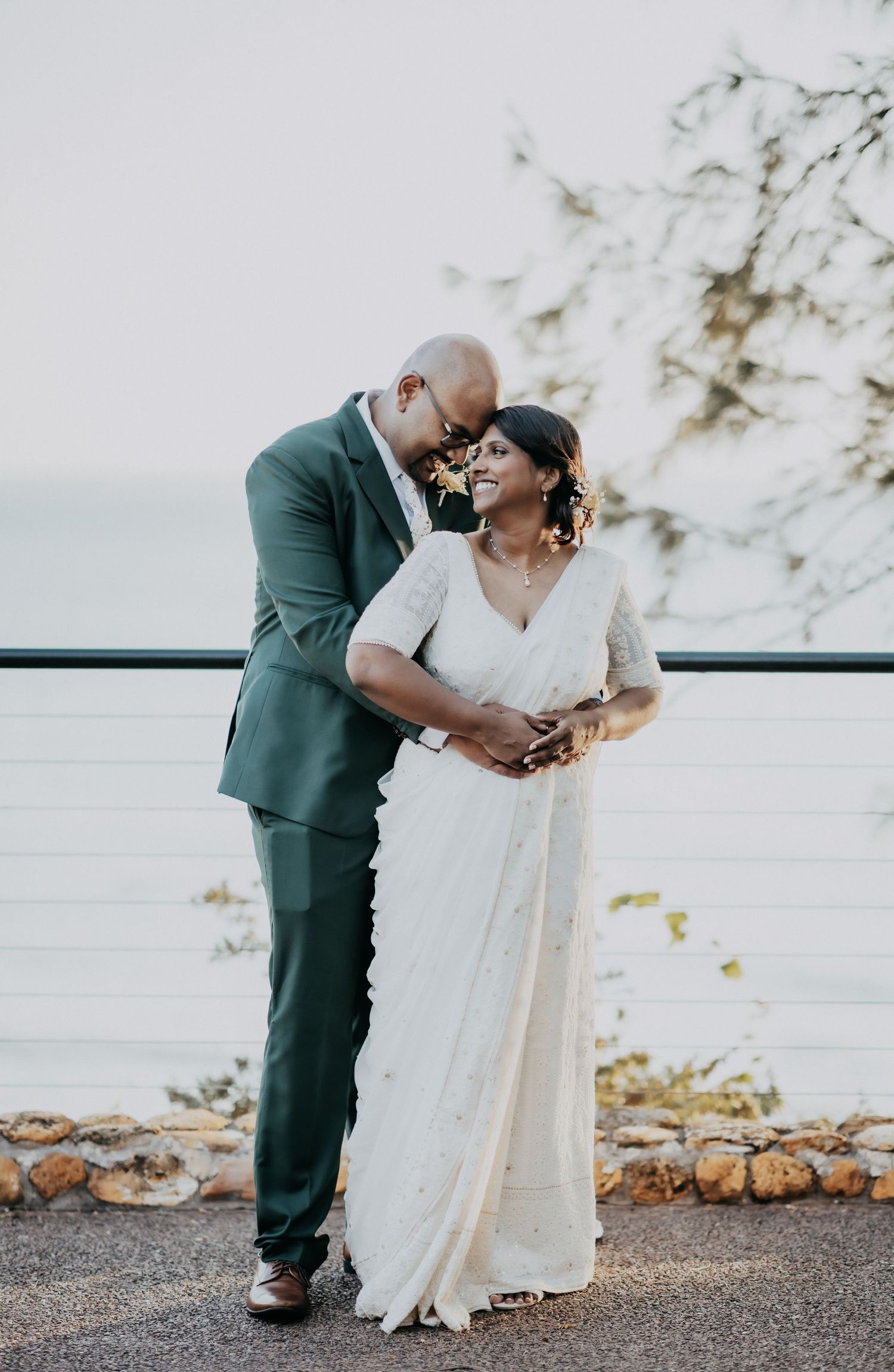 Couple sharing romantic moment outdoors, bride in white embroidered saree, groom in green suit by waterfront.