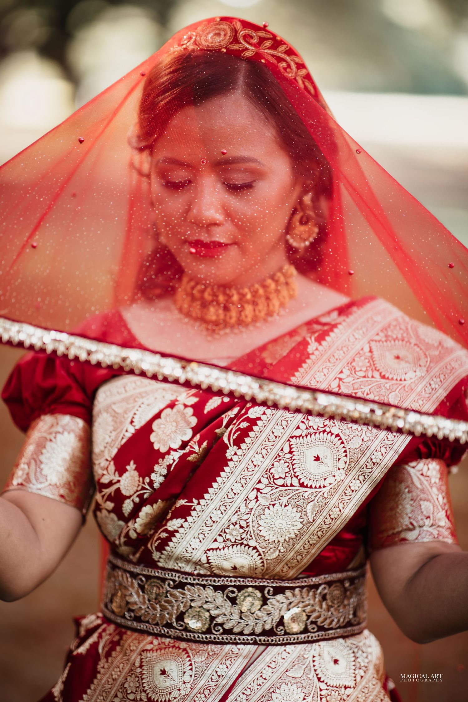 Close-up of Indian bride in red embroidered saree holding red veil overhead with eyes closed.