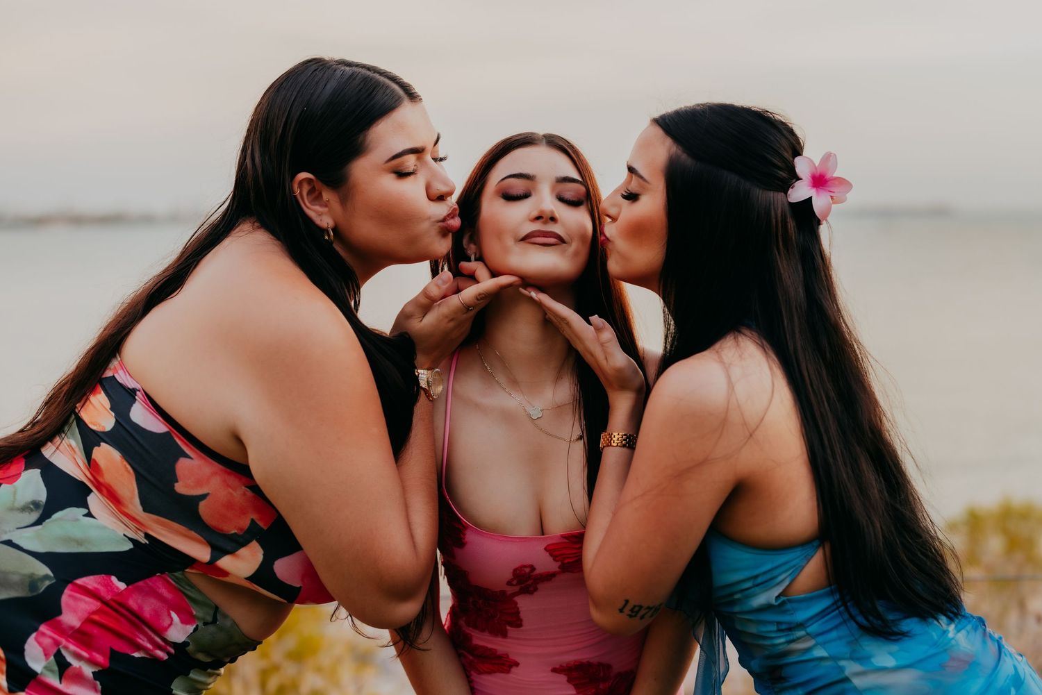 Three young women posing together outdoors, two kissing the cheeks of the woman in the middle.