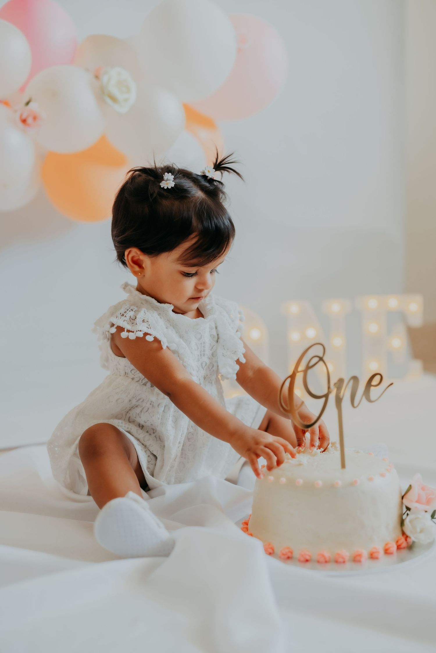 Baby girl in white dress reaching for first birthday cake with gold One topper and peach balloon garland background.