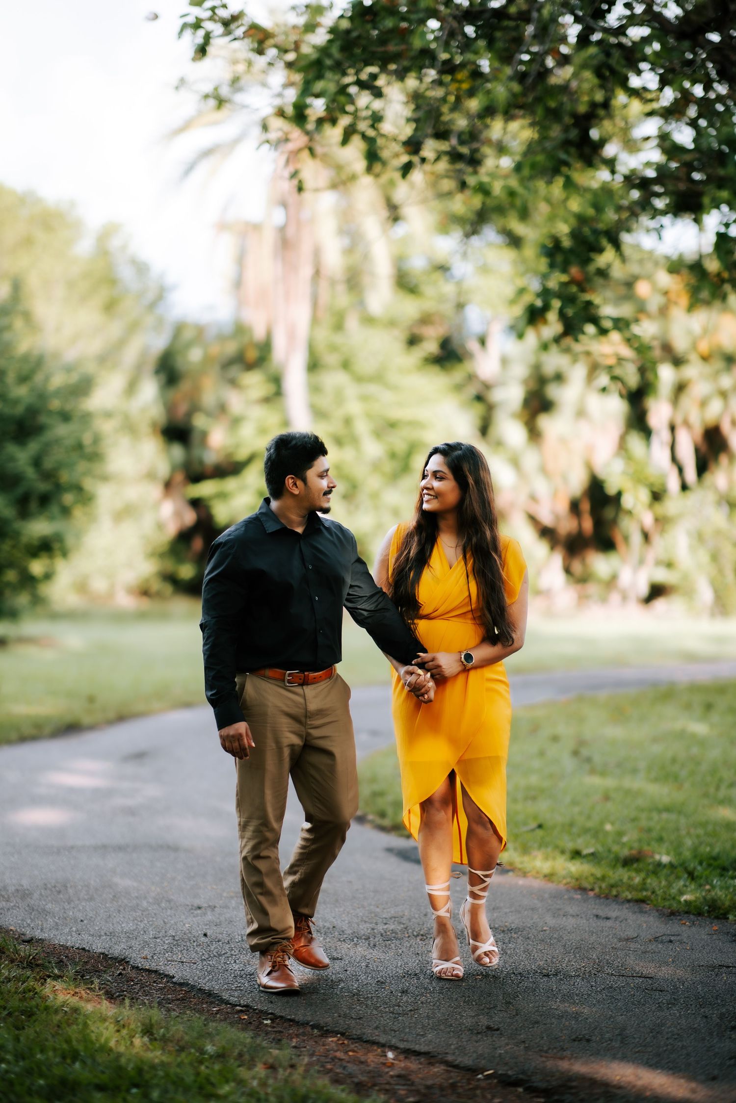 Happy couple walking hand in hand along park path, woman in yellow dress and man in black shirt.