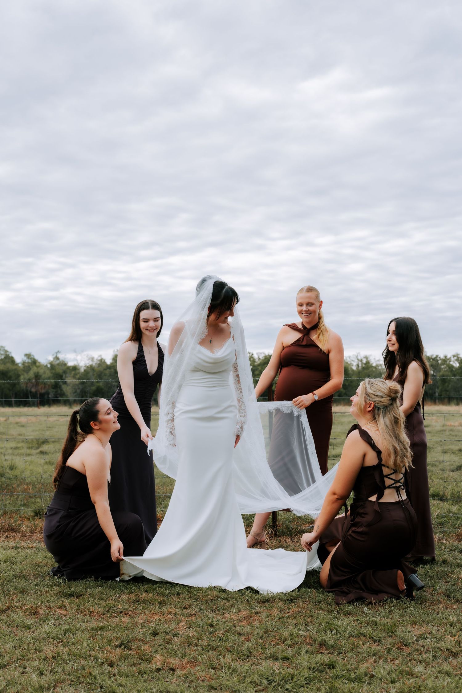 Bride in white gown and veil surrounded by bridesmaids in black and brown dresses on a green field.