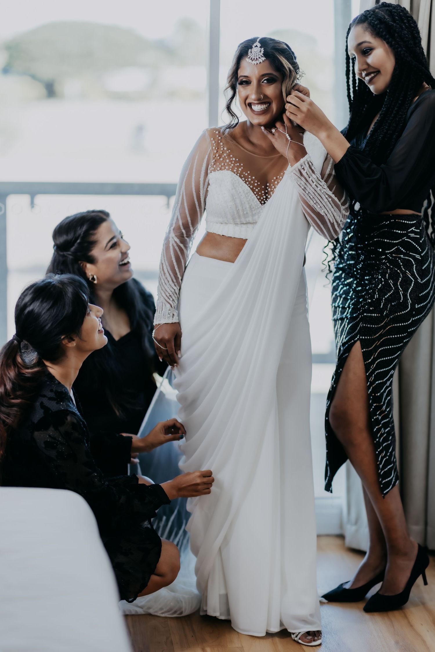 Bride in white beaded saree being helped by bridesmaids while smiling on wedding day.