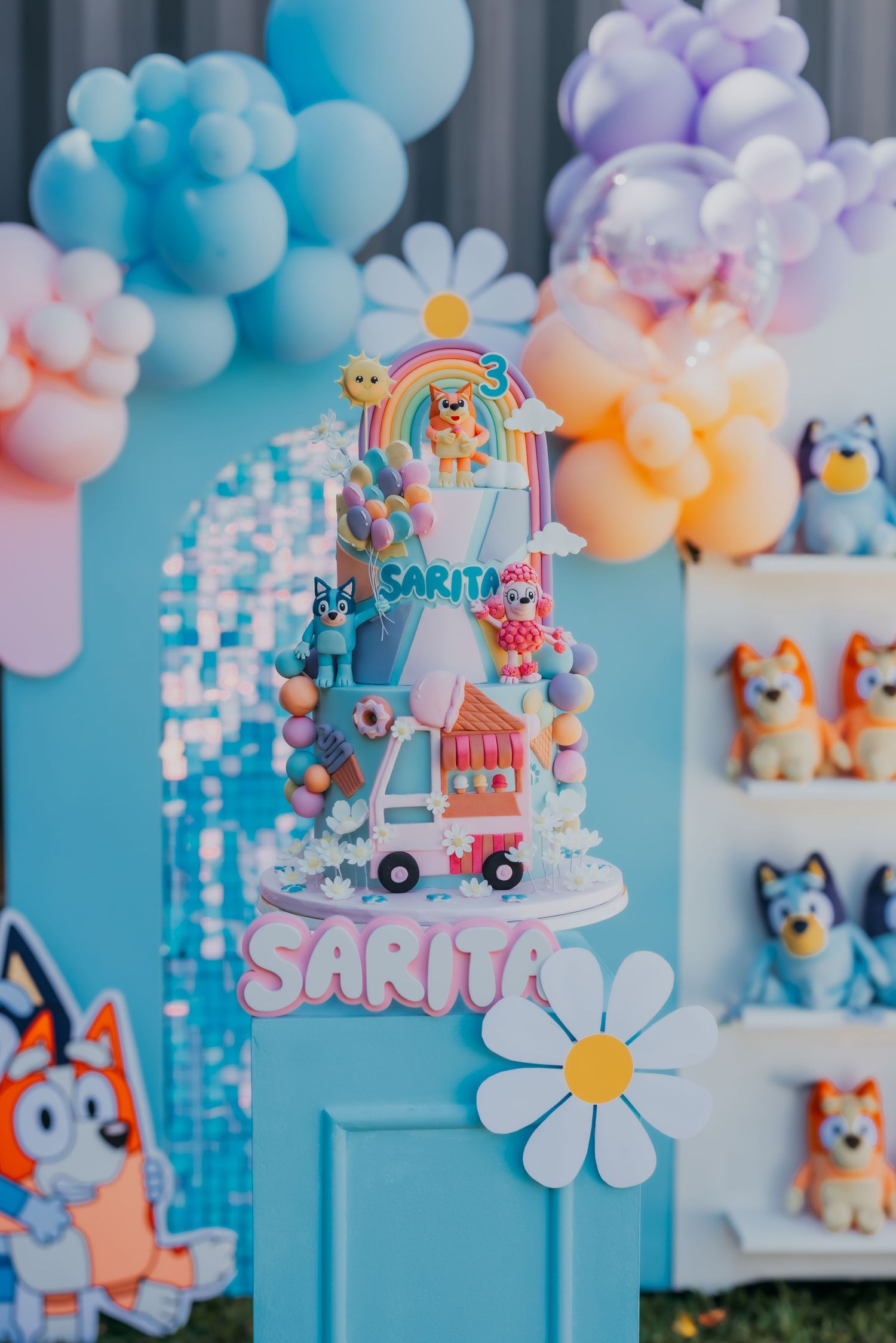 Bluey themed birthday cake with name Sarita displayed on decorated party backdrop.