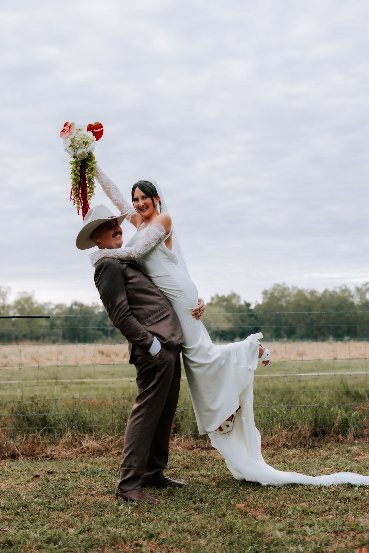 Groom lifting bride in white gown while she raises bouquet in an open field.