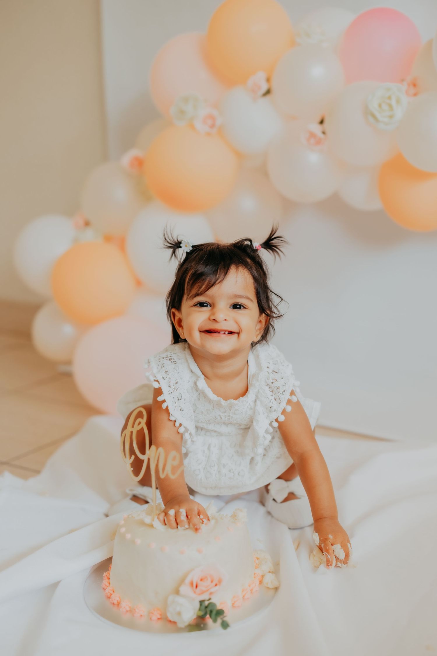 Adorable baby girl in white lace dress smashing first birthday cake with peach and white balloon backdrop.