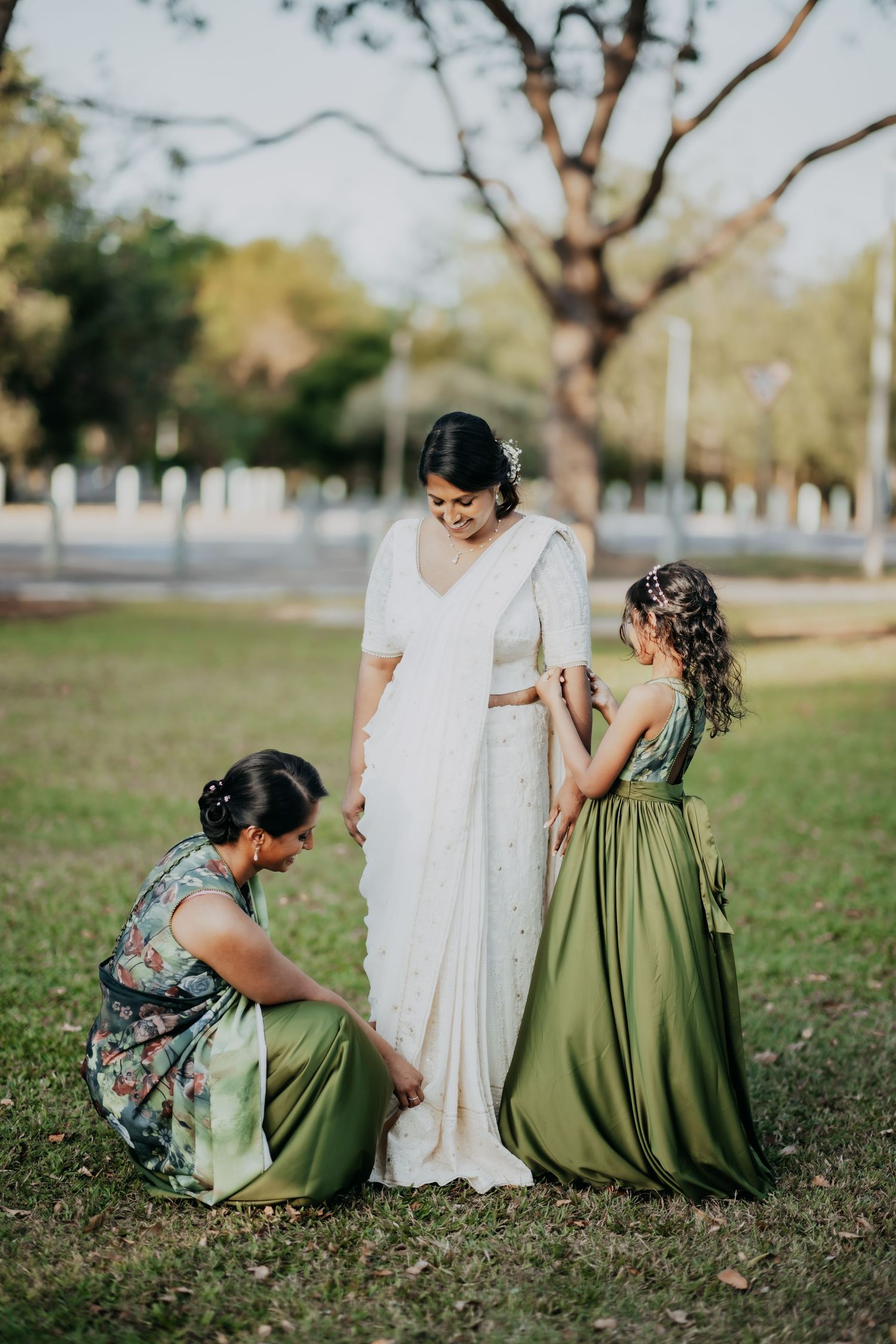 Bridesmaids in green dresses help bride adjust her white saree in a sunlit outdoor park setting.