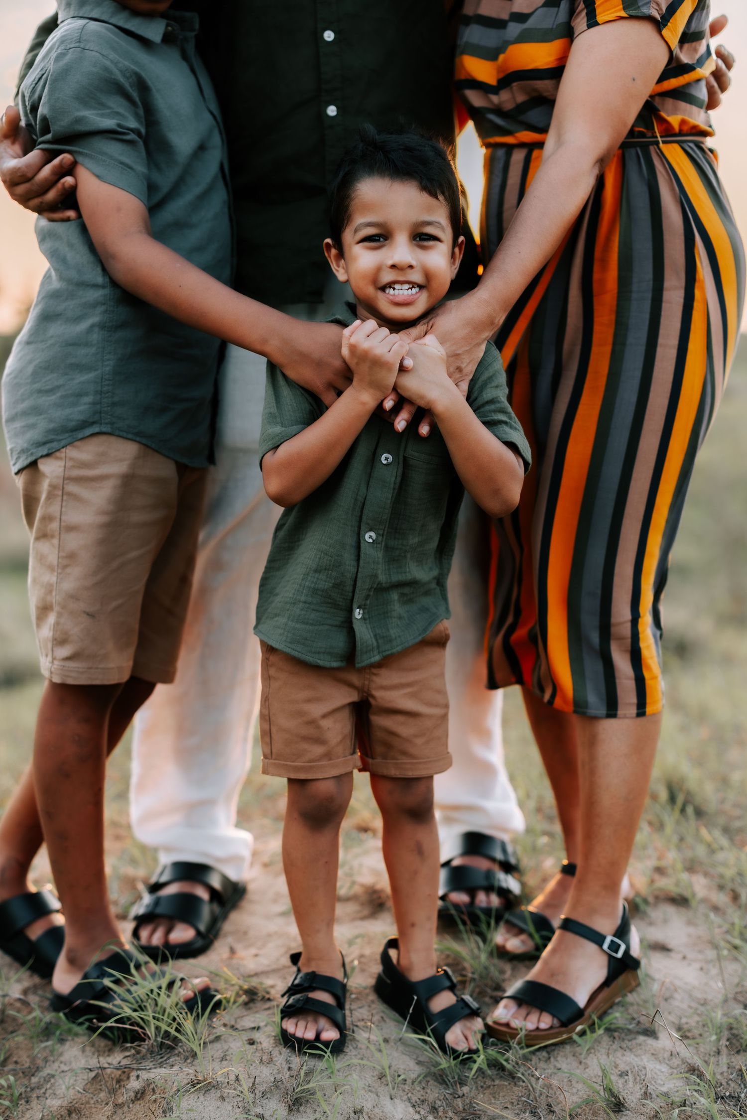 Smiling young boy in green shirt holding parents hands while standing between his family outdoors at sunset.