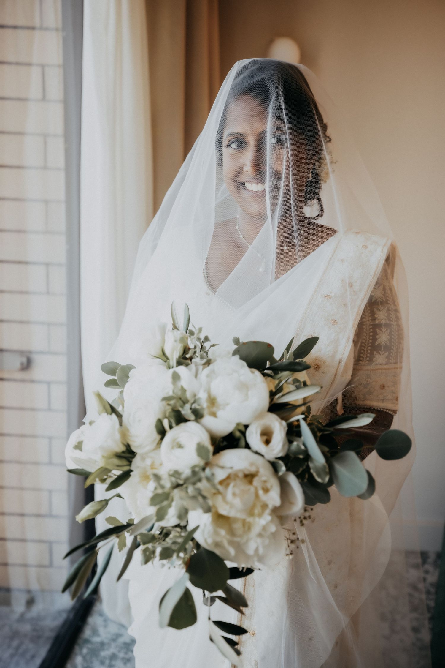 Smiling bride wearing white veil holds large bouquet of white peonies and greenery.