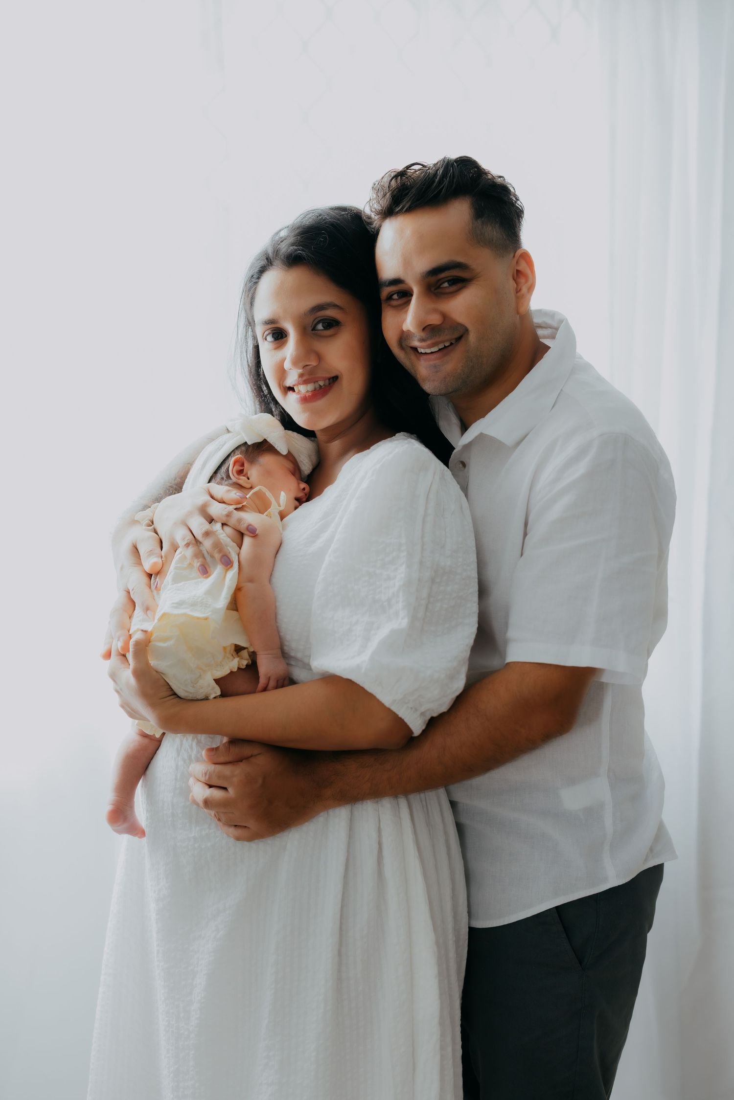 Happy couple in white outfits holding their newborn baby against a bright white background.