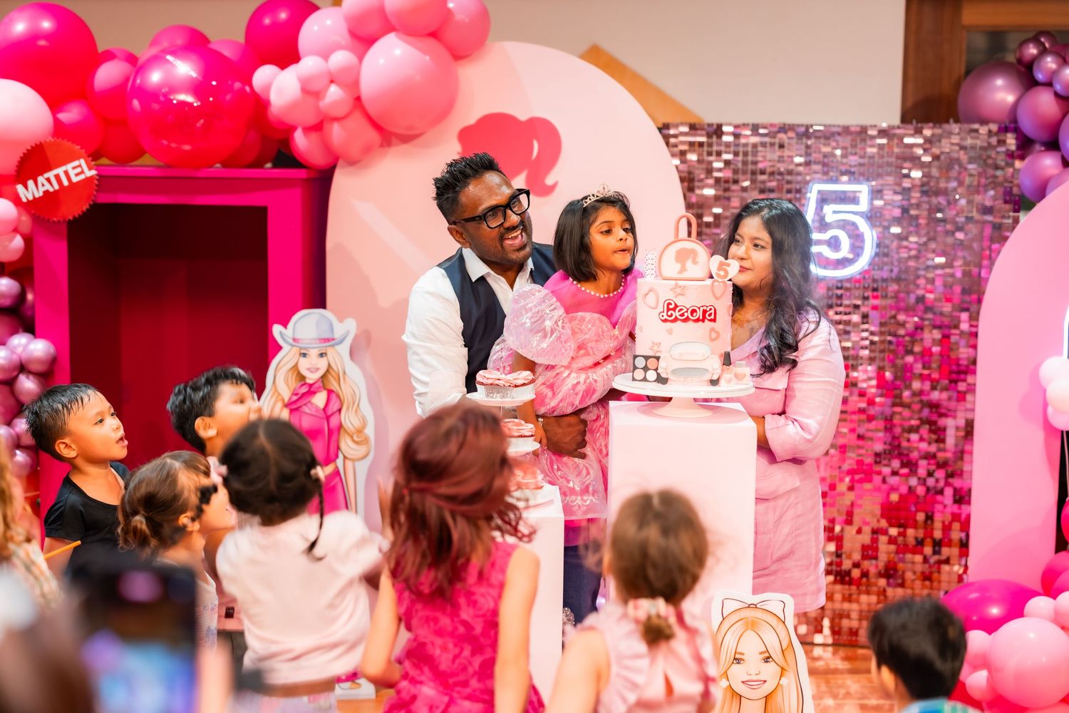 Family celebrating a Barbie themed 5th birthday party with pink balloons, decorations, and birthday cake on stage.