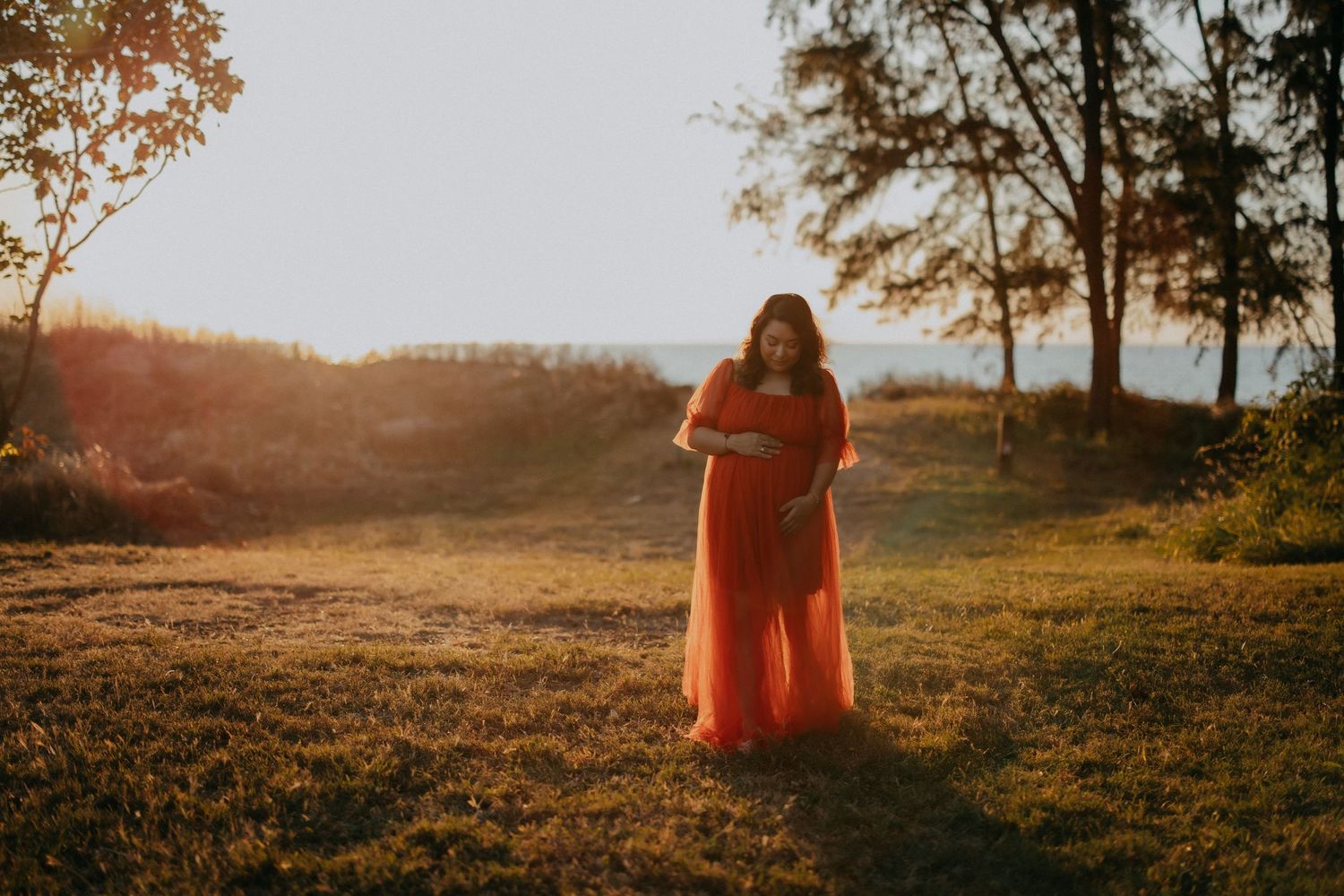 Pregnant woman in flowing orange dress standing in sunlit park at golden hour during maternity shoot.