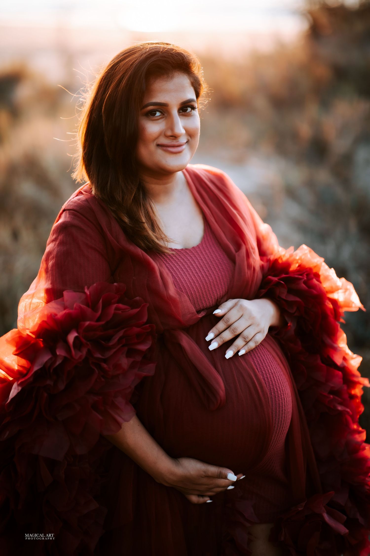 Smiling pregnant woman in burgundy ribbed dress with red ruffled wrap during outdoor photo shoot.