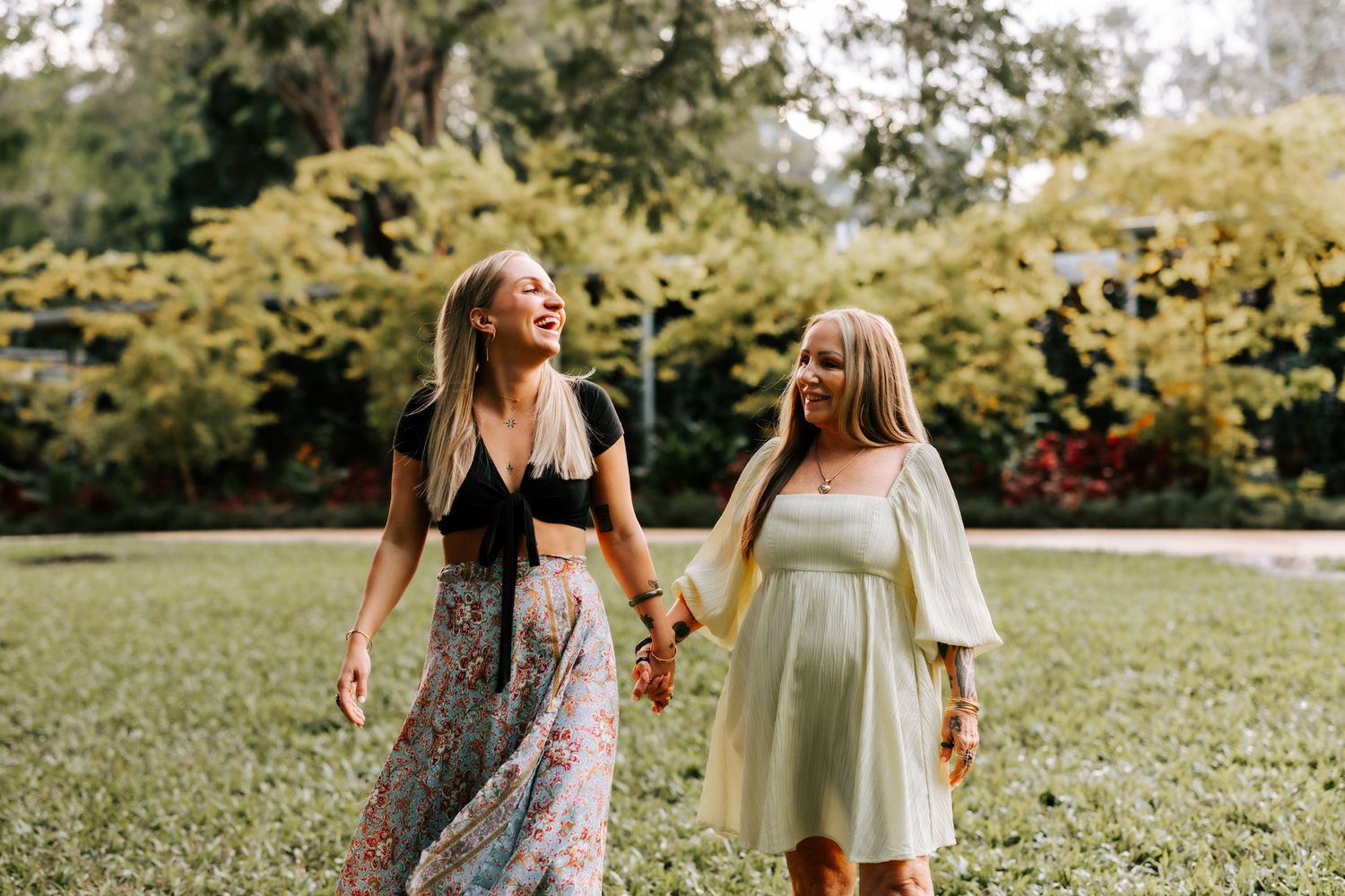 Two women in floral dresses hold hands and laugh while walking through a lush green park.