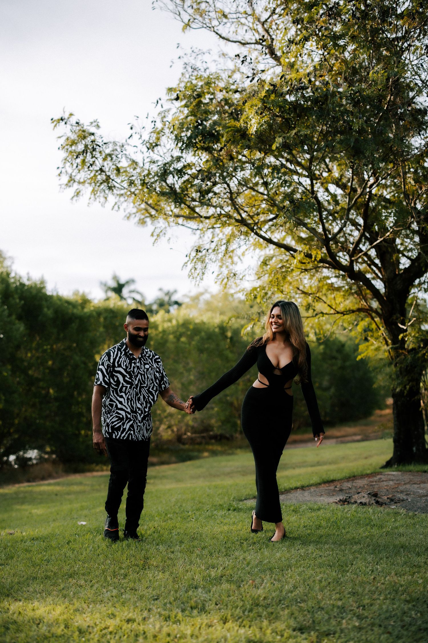 Couple holding hands walking on green grass in a park, man in zebra print shirt, woman in elegant black dress.
