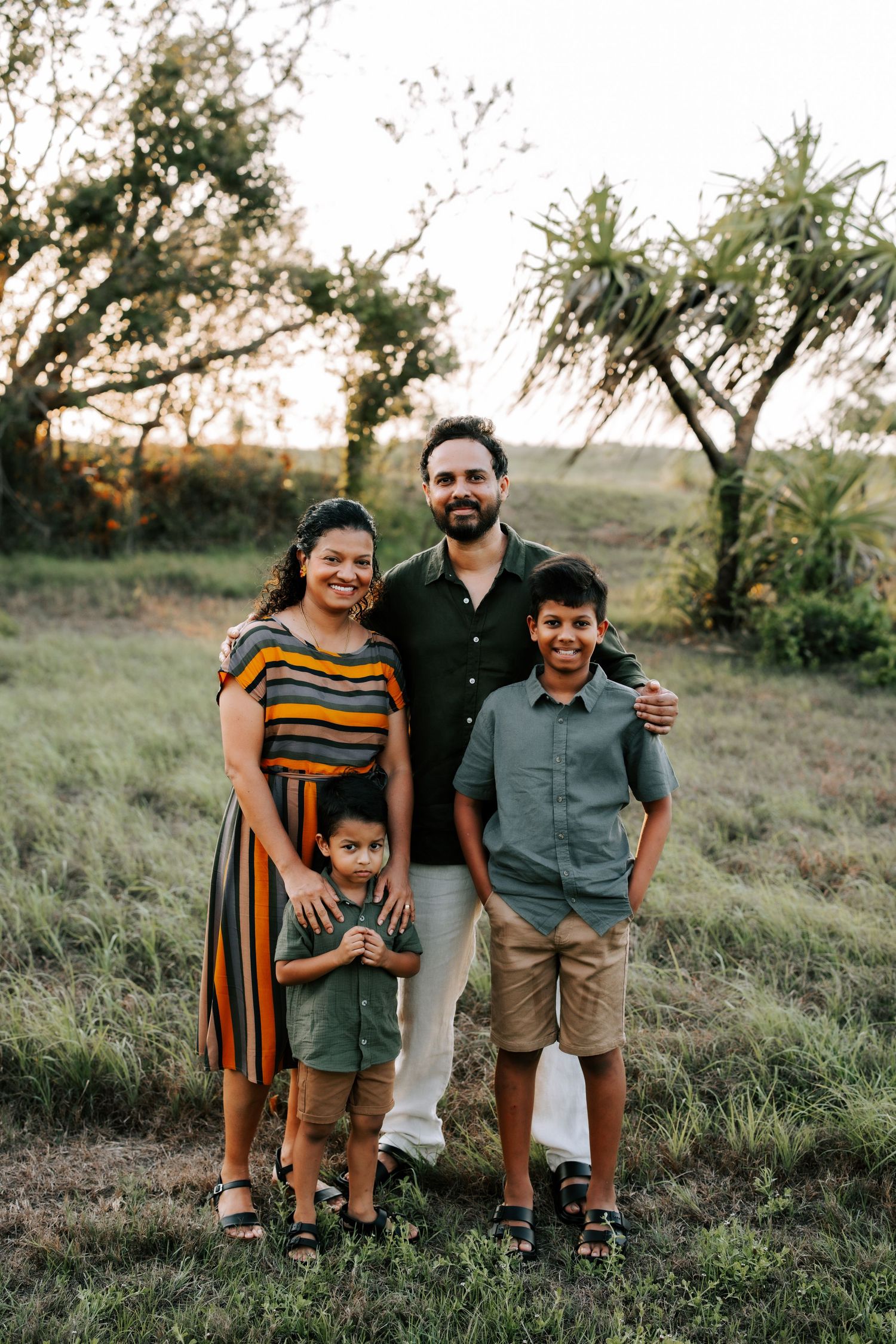 Happy family of four posing outdoors at sunset, parents with two young boys in a grassy field with trees.