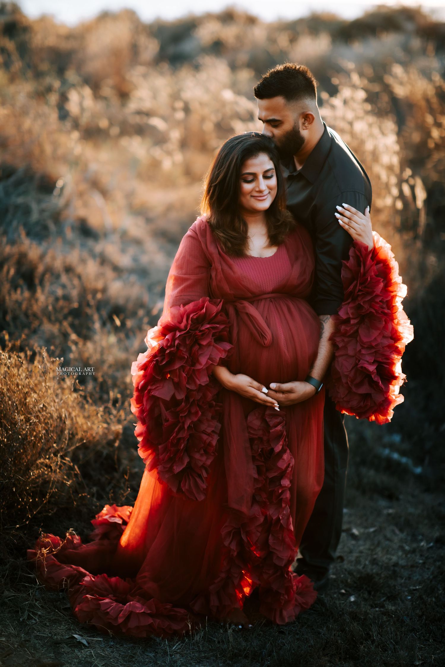 Couple maternity photo shoot with woman in red ruffled gown and man in black suit at sunset.