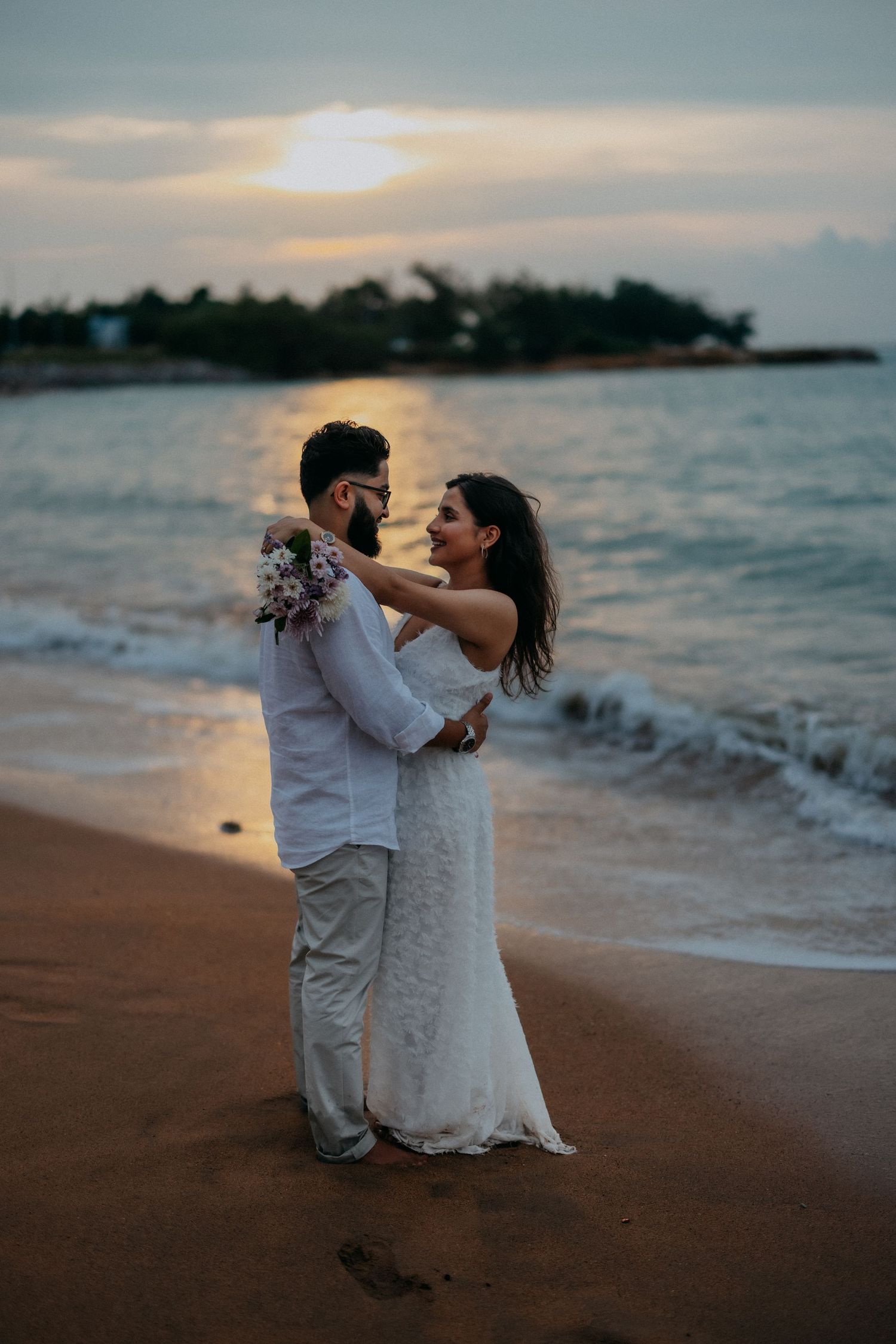 Couple embracing on beach at sunset, bride in white dress holding bouquet with ocean waves behind them.