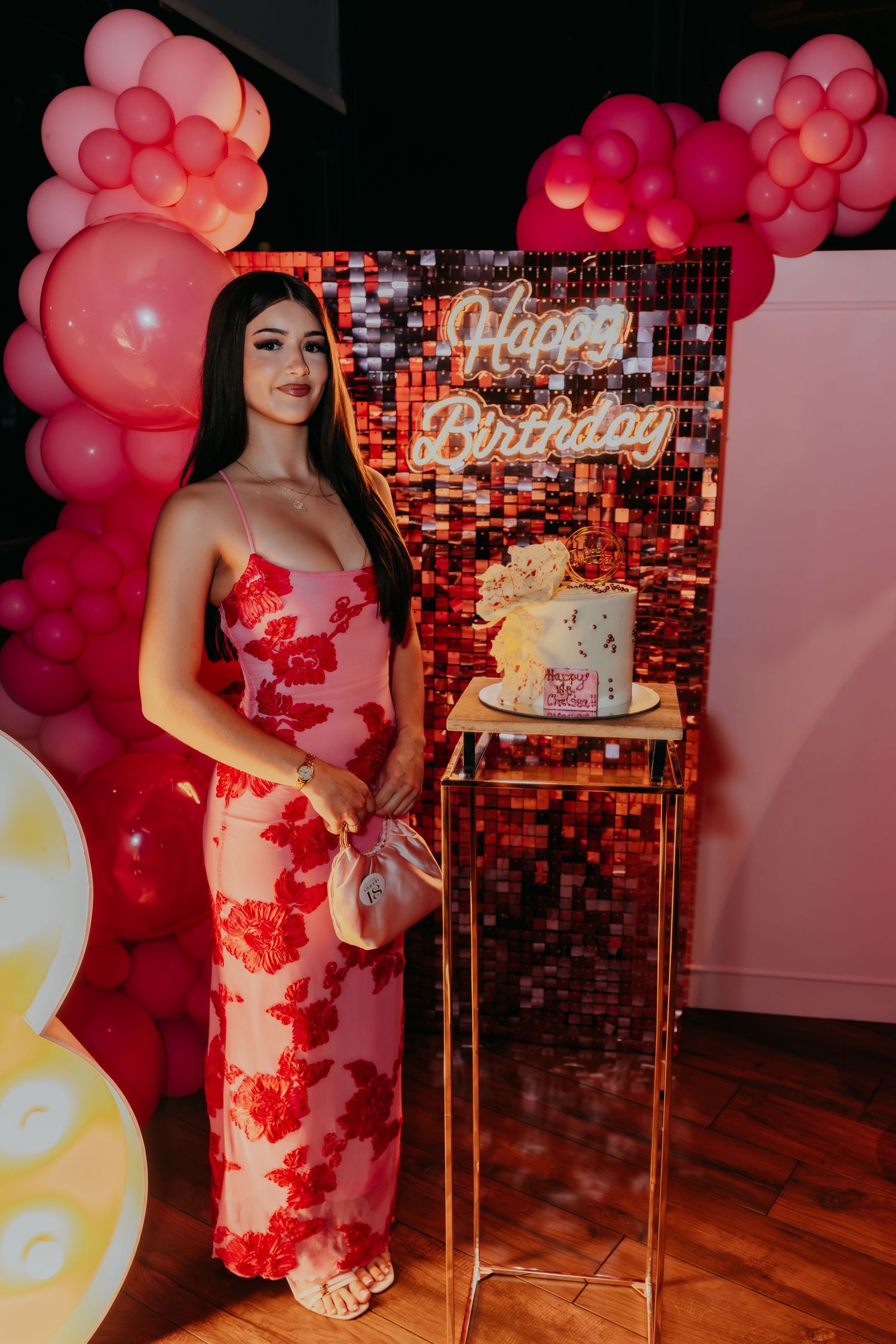 Woman in pink floral gown standing beside birthday cake at decorated party venue.
