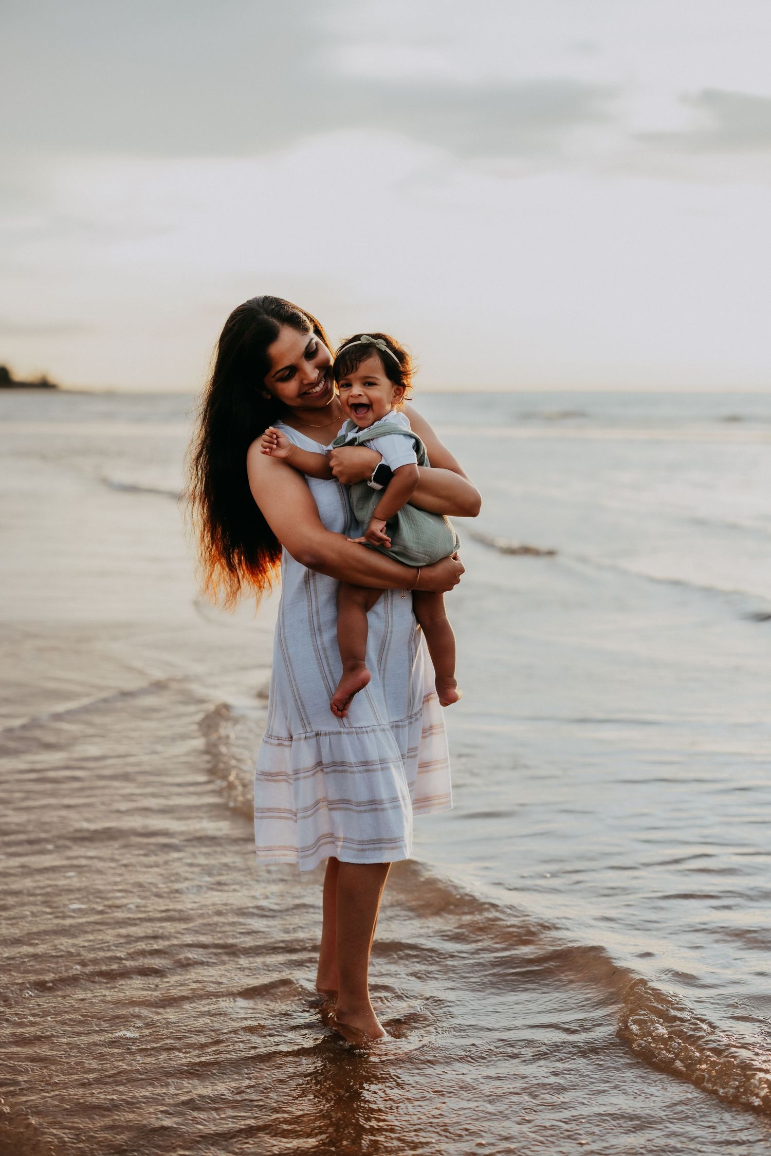 A laughing mother holds her happy baby girl while standing in ocean waves at sunset.