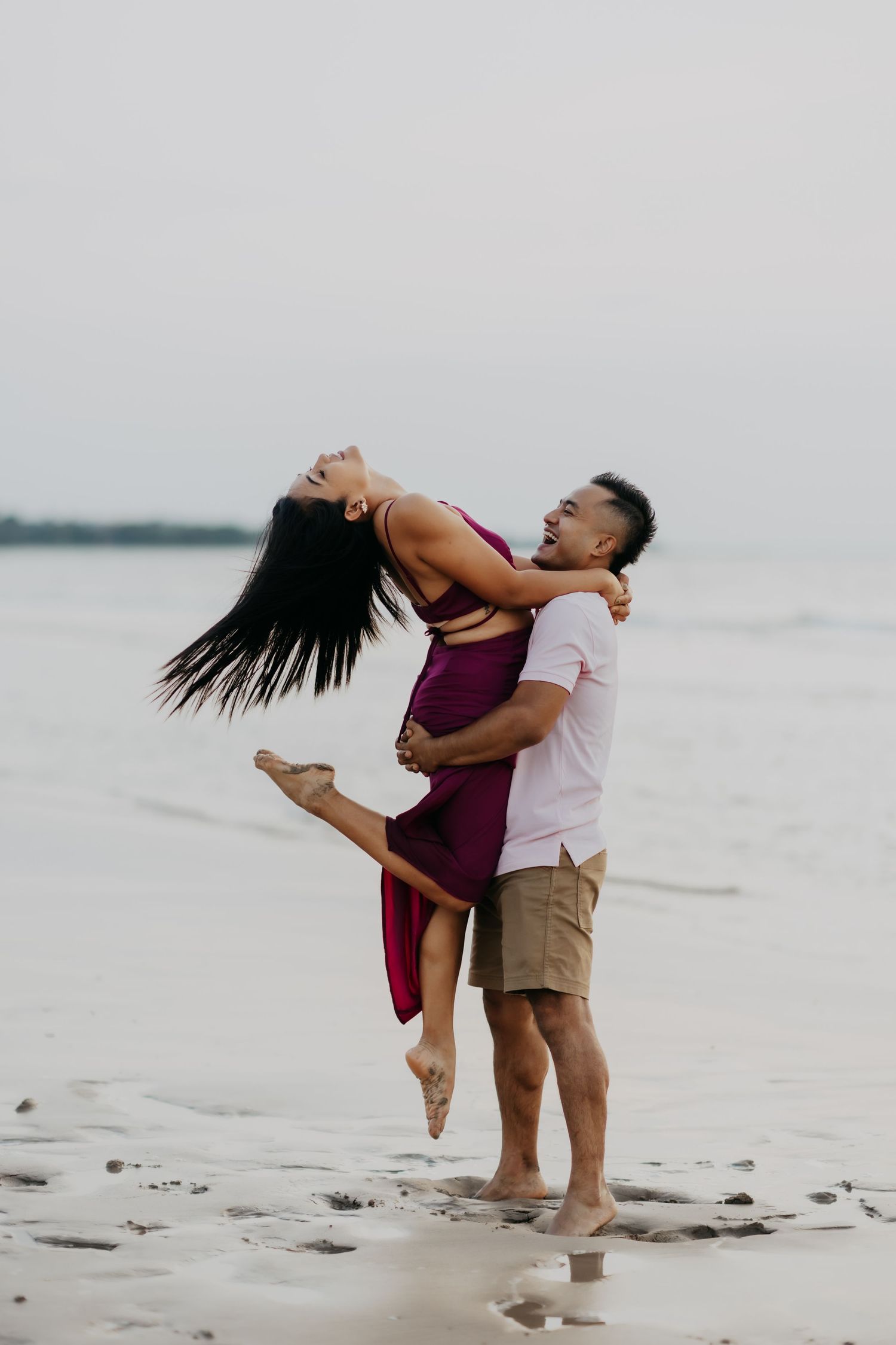 A happy couple embraces on a beach, the man lifting the laughing woman in a burgundy dress.