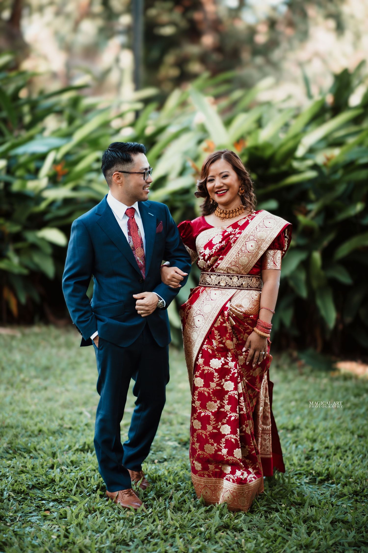 Couple smiling at each other outdoors, bride in red gold saree, groom in navy suit.